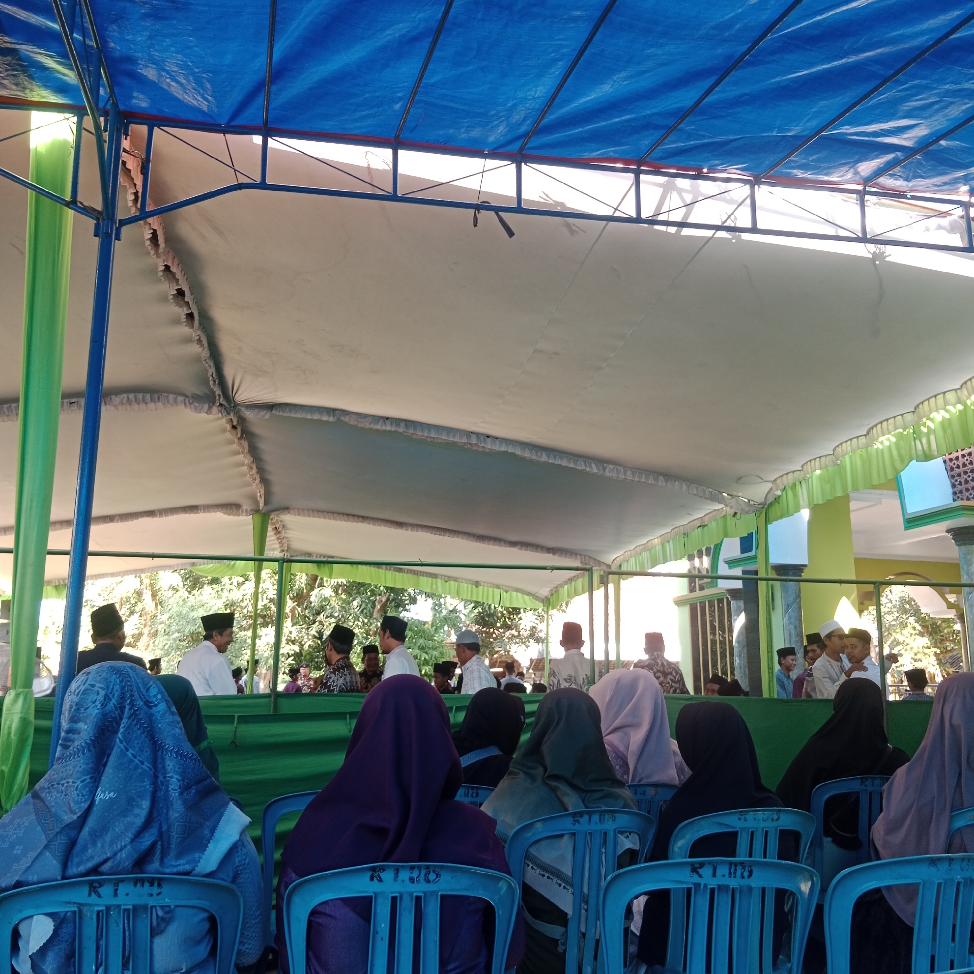 The image depicts an outdoor gathering under a blue tarp shelter, possibly a temporary structure erected for an event. The shelter is supported by blue poles and green fabric drapery. In the foreground, several people, primarily women wearing hijabs in various colors including blue, purple, and black, are seated on blue plastic chairs. The chairs have markings on their backs. Behind them, a group of men, some wearing peci hats, are standing and conversing. The background reveals lush green trees and foliage, suggesting a semi-rural or garden setting. To the right, a building with green and yellow accents, possibly a mosque or community hall, is visible, along with decorative pillars. The lighting indicates it is daytime, likely sunny, casting shadows. The presence of people in traditional attire and the building's architecture suggest a cultural or religious event taking place in Tegowanu Kulon, Indonesia.