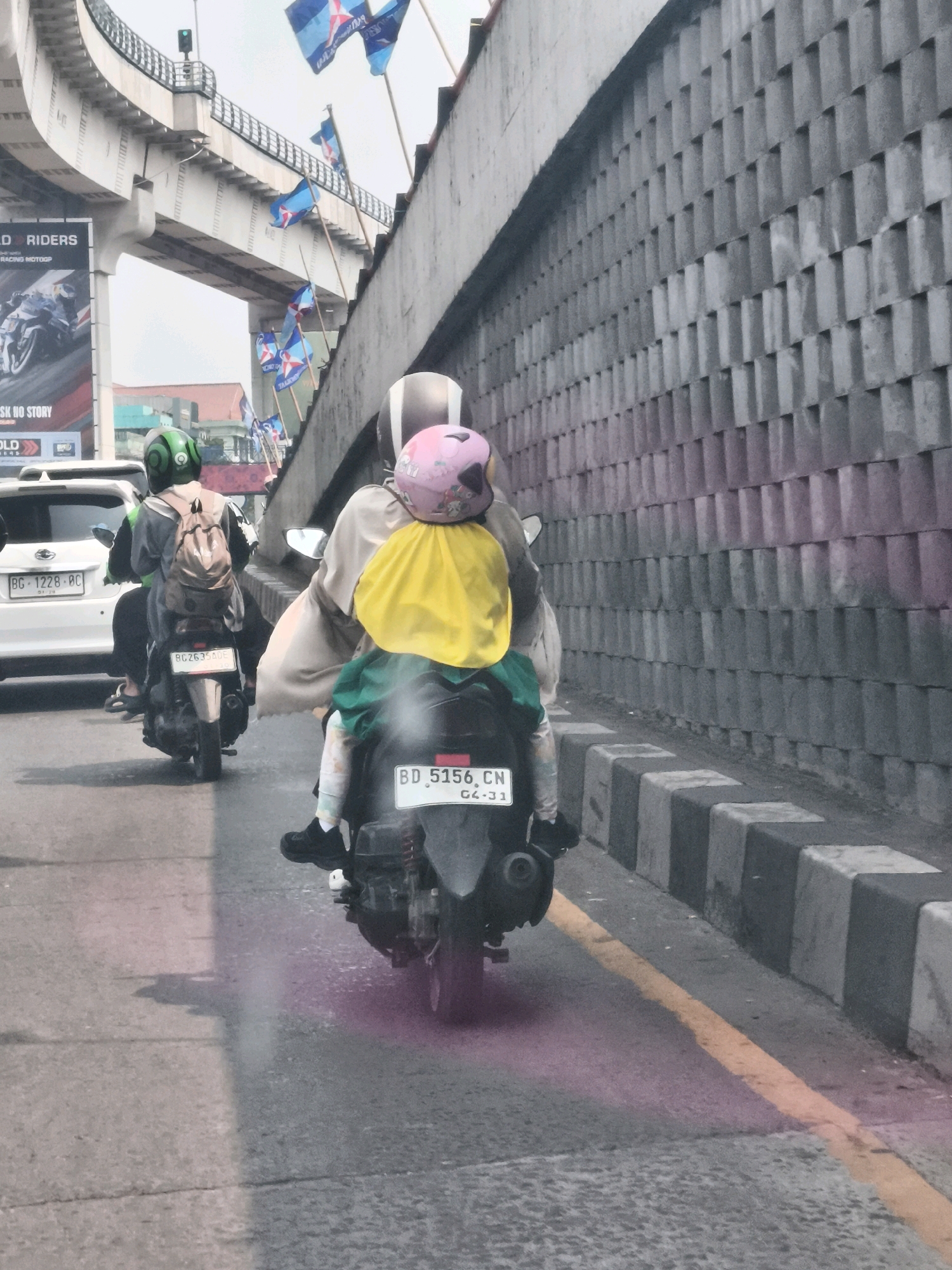 In Palembang, Indonesia, on a cloudy day, a person wearing a green helmet and a backpack rides a motorcycle. They are followed by another motorcycle carrying two people. The person in the front has on a light-colored robe and is wearing a pink helmet with a yellow cloth draped over it. The person behind them is wearing a green garment. Both motorcycles have license plates. The scene is on a street with a concrete wall on the right and an elevated road structure on the left. Several flags are visible attached to poles along the elevated road. The road has a yellow line marking the lane.