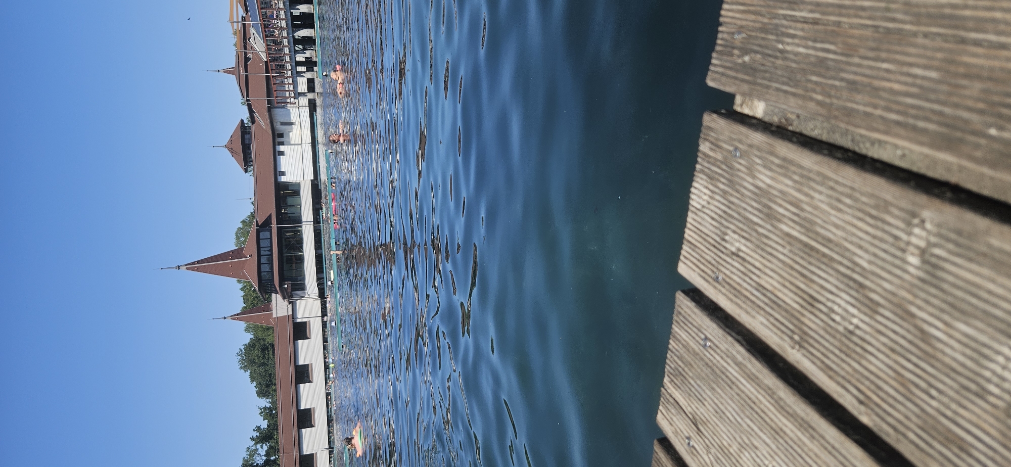 The image depicts a sunny outdoor scene at the Hévíz Thermal Lake in Hévíz, Hungary. In the foreground, weathered wooden planks of a pier are visible, leading into the tranquil, deep blue water of the lake. The water's surface is gently rippled, reflecting the clear blue sky.
In the middle ground and background, the iconic architecture of the lake's bathhouse complex is prominent. The buildings feature reddish-brown conical roofs with spires, typical of Hungarian architectural styles, and are painted white. The structure extends over the water, with walkways and covered areas visible.
Several individuals are enjoying the lake. Some are floating on inflatable rings, including a person in a green ring in the left middle ground, and others are lounging on pink inflatable mats further in the distance. The people appear relaxed and are engaged in leisurely aquatic activities.
The overall environment is serene and suggests a warm, pleasant day at a popular outdoor thermal spa. Trees and greenery can be seen behind the bathhouse, contributing to the natural setting. The clear sky indicates good weather conditions.