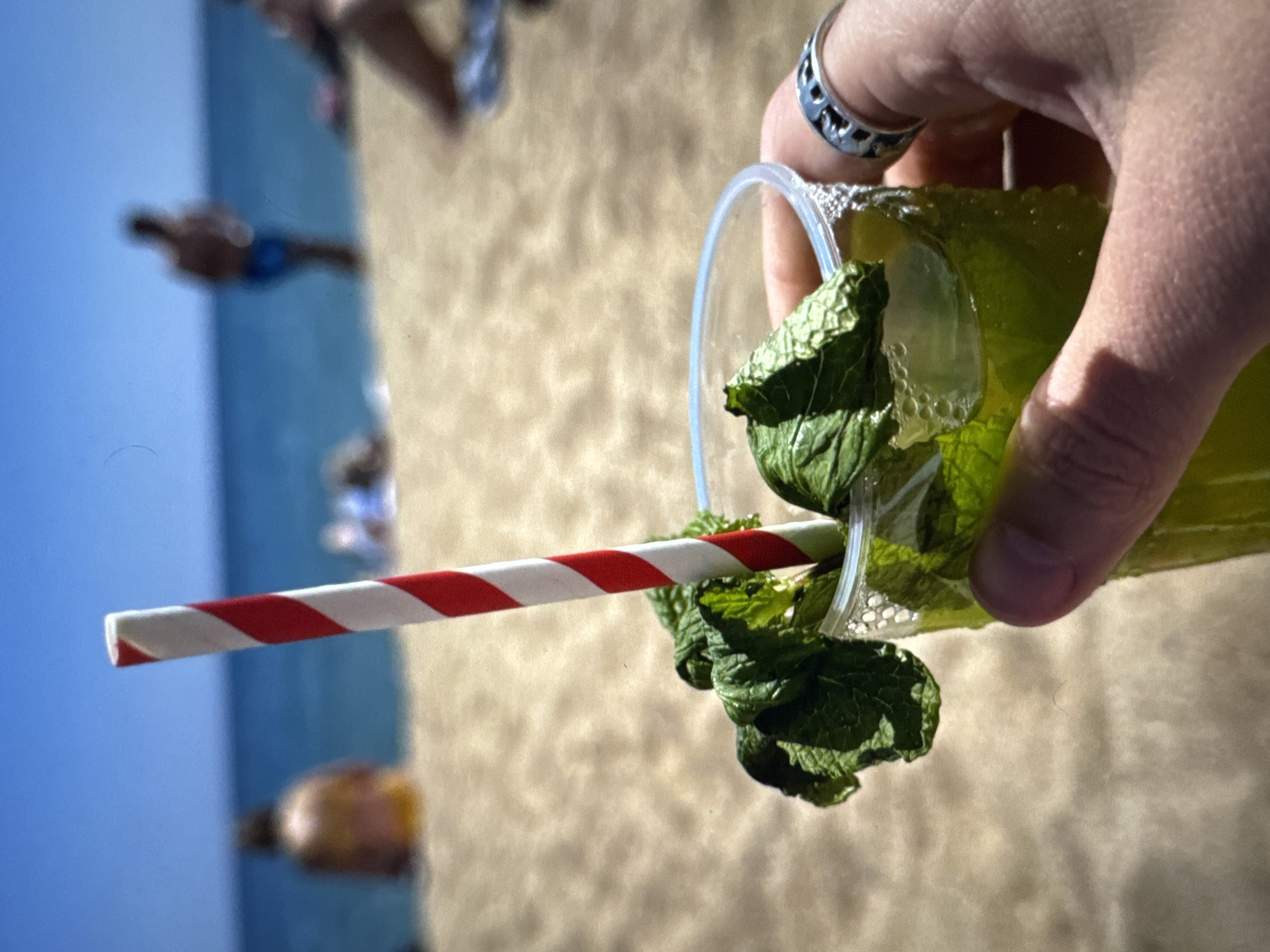 In the foreground, a person's hand, adorned with a silver ring, holds a clear plastic cup filled with a green beverage. The drink is garnished with fresh mint leaves and contains ice cubes. A red and white striped straw protrudes from the cup. The background depicts a sandy beach with the blue ocean and clear sky beyond. Several blurred figures are visible on the beach and in the water, suggesting a summer day at a seaside location. The image does not depict any elements specific to the city of Porrentruy, Switzerland, as it is an outdoor beach scene.