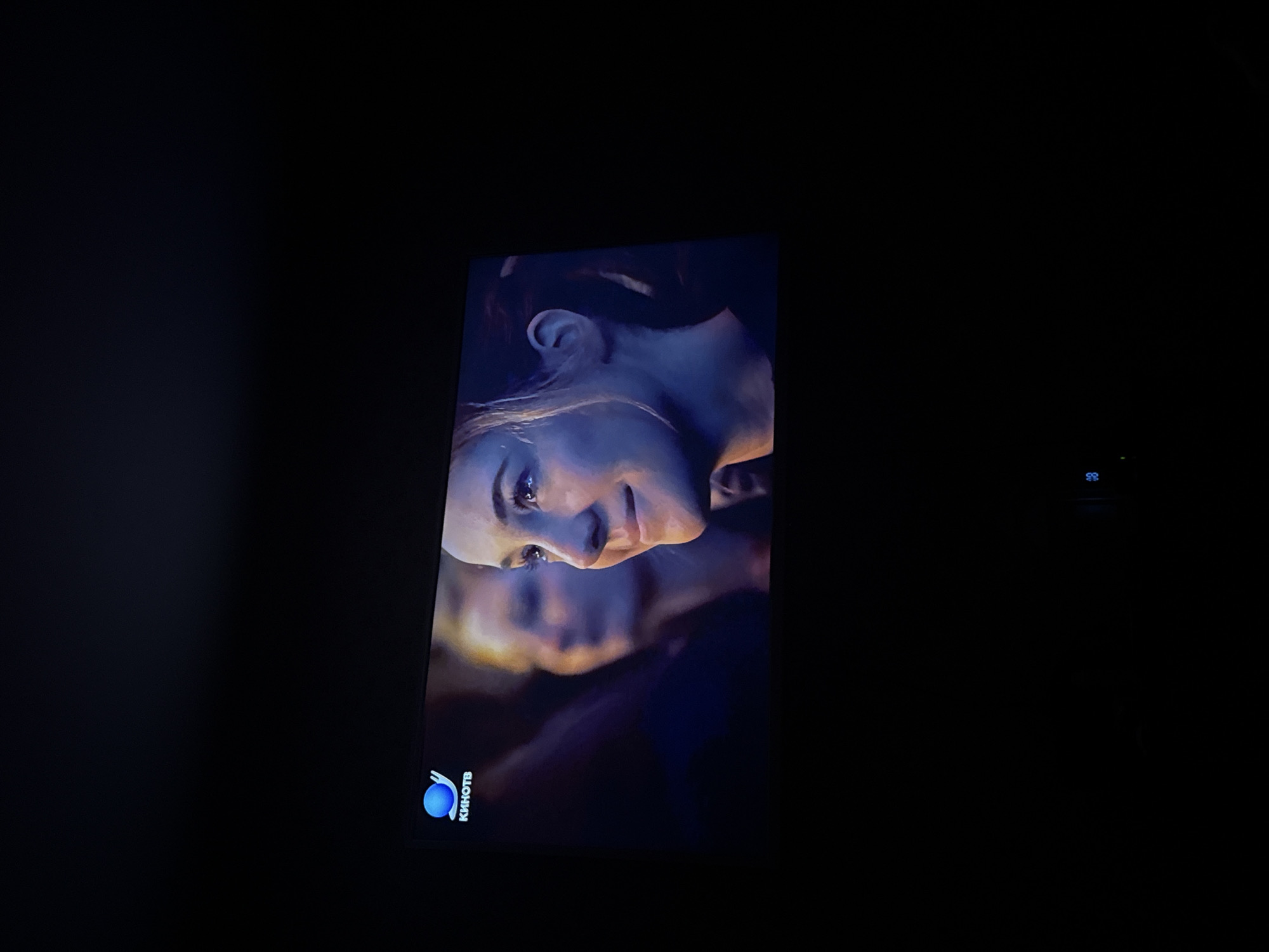 The image shows a close-up of a television screen displaying a scene with two people. The main subject is a woman with light skin and visible eyelashes, tilted at an angle. A second person is blurred in the background behind her. In the bottom left corner of the screen is a blue and white logo with the word "KHHOTB" next to it. The screen is surrounded by darkness. The image does not appear to contain information that allows determining the specific location in Mytishchi, Russia.