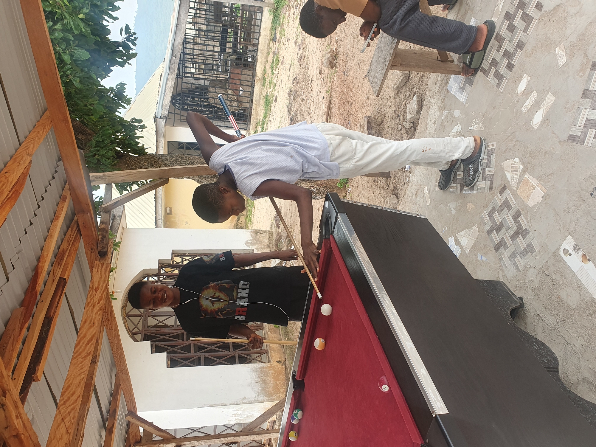 In Lokoja, Nigeria, two young men are engaged in a game of billiards outdoors under a rustic, open-sided structure. One man, wearing a black t-shirt with a graphic design and headphones, stands at the far side of the pool table, holding a cue stick. The other, in a light blue sleeveless shirt and off-white pants, is actively playing, aiming a shot with his cue. Several billiard balls are scattered on the red felt of the table. To the right, a third individual is seated on a wooden bench, looking down at a mobile phone. The surrounding environment appears to be a dusty, open area with some vegetation and buildings in the background, characteristic of a less developed urban setting. The ground is a mix of concrete tiles and sand.