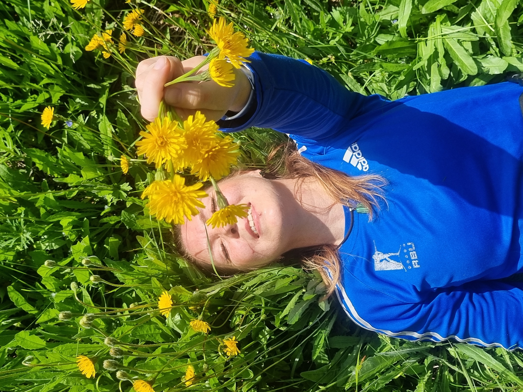 A young woman with reddish-brown hair lies on her back in a vibrant green field, surrounded by numerous bright yellow dandelions. She is wearing a blue long-sleeved athletic shirt. Her head is tilted slightly, and she is smiling broadly with her eyes squinting, suggesting she is enjoying the bright sunlight. She holds a cluster of dandelions in her right hand, positioning them near her face.
The scene is illuminated by strong, clear daylight, indicating a sunny day. Dandelions are abundant across the field, some in full bloom and others as closed buds. The woman's blue shirt features a white "adidas" logo on the left upper chest/sleeve and another white logo with a stylized figure and the text "RBU" on the right chest area. The overall impression is one of joy, relaxation, and a connection with nature in a sunny meadow. The location context is Chaikovsky, Russia.