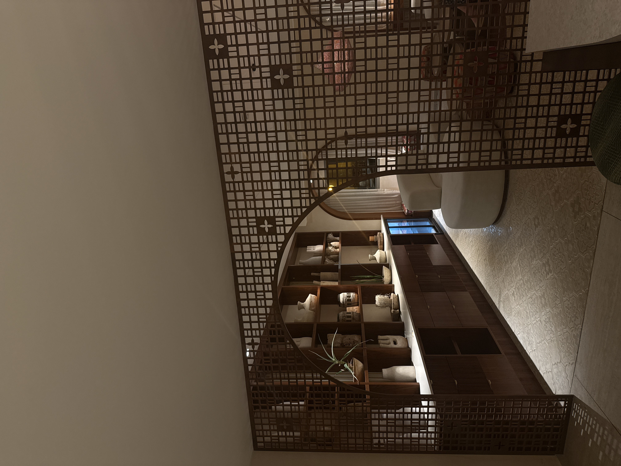 This is an indoor scene of a modern living room or lounge area, possibly in Tamar Regional Council, Israel, featuring an arched doorway and an intricate wooden screen divider. The room is dimly lit, with warm lighting highlighting the textures and objects.
In the foreground, a decorative wooden screen with a geometric lattice pattern partially obstructs the view. Through this screen, an arched doorway leads to another part of the room.
To the left, a tall, built-in wooden shelving unit is filled with various decorative vases, pottery, and plants. The shelves are organized with a minimalist aesthetic, displaying items of different shapes, sizes, and textures, predominantly in earthy tones. Below the shelves, a series of wooden drawers and cabinets form a long sideboard.
To the right of the arched doorway, a plush, white, circular sofa is visible. Further back, through the arch and past the sofa, elements of another seating area can be glimpsed, including a patterned rug and what appears to be a woven decorative lamp with red accents. The floor throughout the visible area is tiled with a light-colored pattern. The walls appear to be a neutral, warm tone. No people are present in the image.