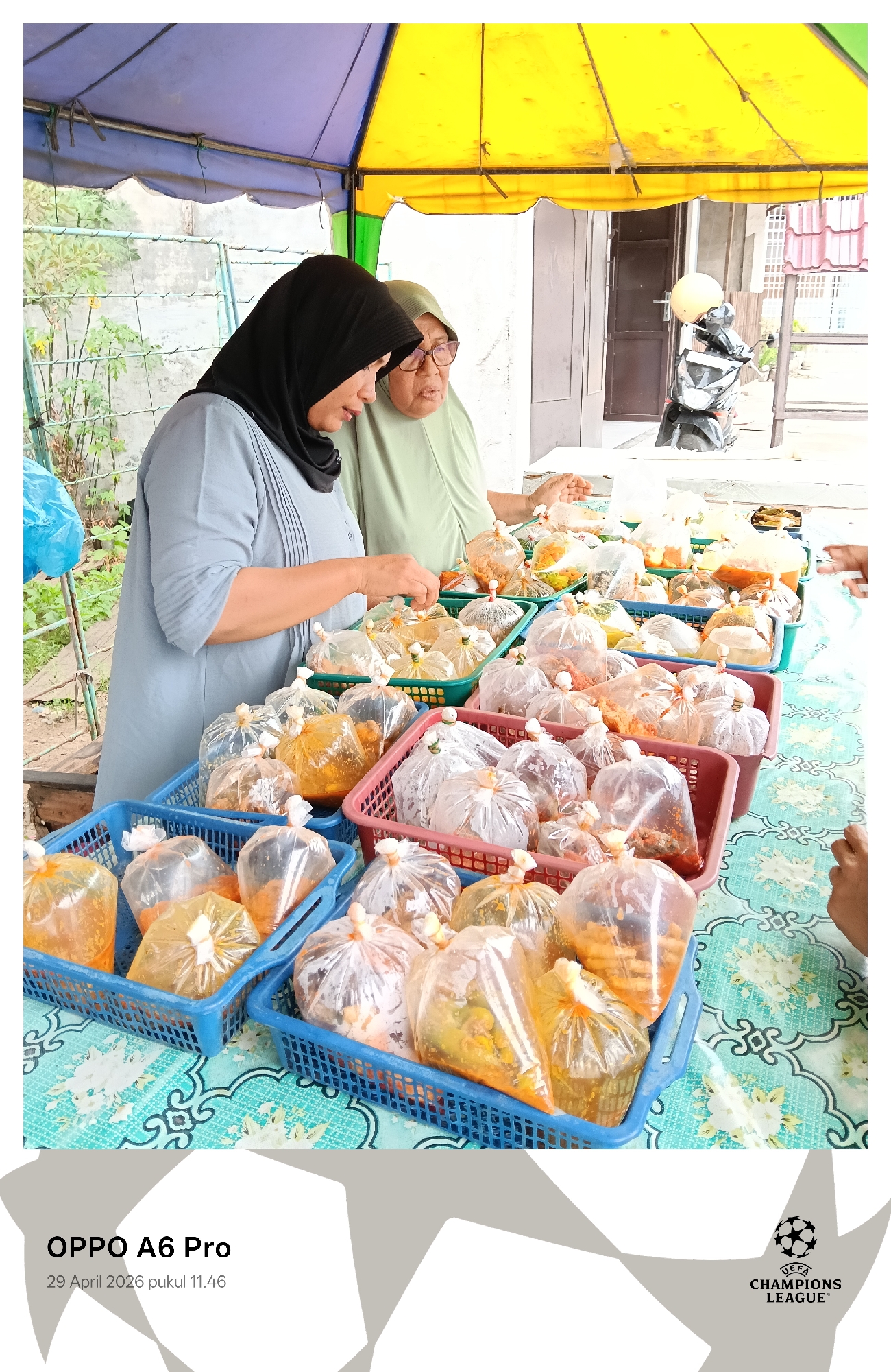 The image captures an outdoor food stall scene in Lhokseumawe, Indonesia, under a blue and yellow canopy. Two women, presumably vendors, are central to the composition. The woman on the left wears a black hijab and a light blue tunic, focused on arranging or tending to the merchandise on the table. The woman behind her, to the right, wears a light green hijab and matching top, looking towards the right, possibly engaging with someone off-camera. Partially visible hands on the far right suggest the presence of other individuals or customers.
The table is covered with a light blue tablecloth featuring a white floral pattern and is filled with numerous plastic baskets. These baskets hold dozens of clear plastic bags, each knotted at the top and containing individual portions of pre-packaged food. The contents appear varied, with some bags showing yellow or orange-tinted dishes, possibly curries or sauces, while others contain solid food items that could be rice, meat, or vegetables, prepared for takeaway.
In the background, a motor scooter with a light-colored helmet parked on it is visible near a building, suggesting a street or residential area where the stall is set up. The bright lighting indicates it is daytime, confirmed by the visible timestamp.
Notable text includes "OPPO A6 Pro" and "29 April 2026 pukul 11.46" printed on the bottom left, indicating the device and time the photo was taken. The "UEFA CHAMPIONS LEAGUE" logo is also present in the bottom right corner. The scene depicts a typical daily commerce activity, likely a street food vendor selling prepared meals.