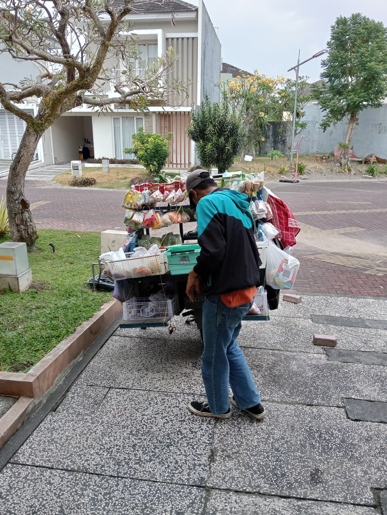 A man in a turquoise and black hooded jacket and blue jeans stands next to a mobile food cart laden with various packaged goods. The cart has multiple shelves displaying items like snacks, drinks, and other food products. The man is positioned on a paved sidewalk in front of a residential area, with a neatly manicured lawn to his left. In the background, modern houses with white facades and prominent windows are visible. A large tree with gnarled branches stands prominently in the foreground, casting a shadow on the scene. The environment suggests a suburban setting in Ngaglik, Indonesia.
