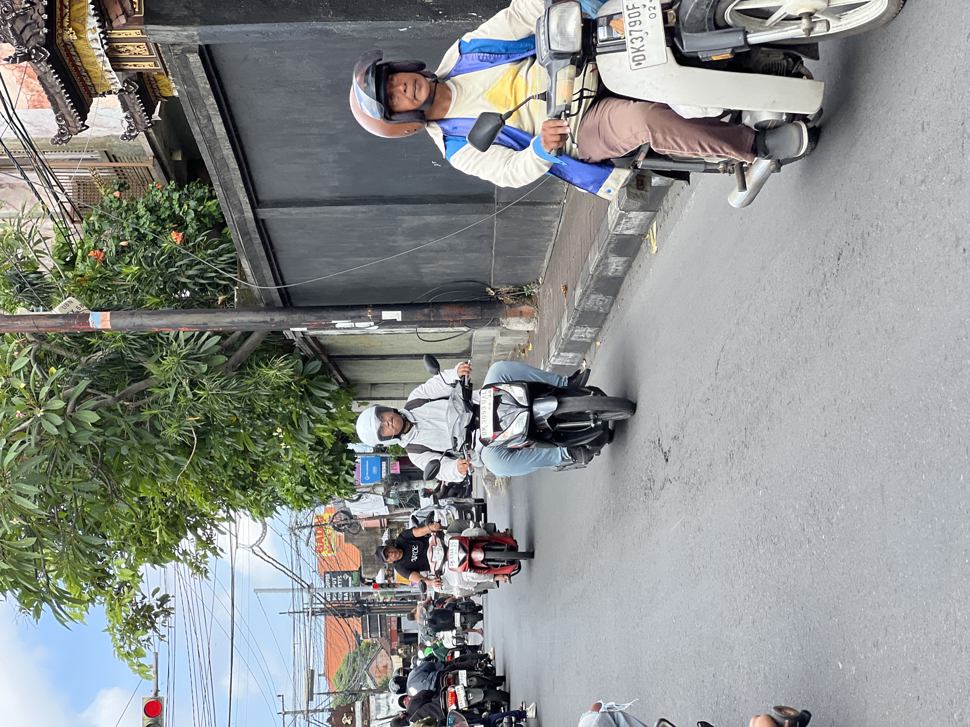 In North Kuta, Indonesia, traffic moves along a paved road lined with a dark grey wall on the right and buildings and lush vegetation on the left. A traffic light with a red signal is visible in the distance. A procession of motorcycles navigates the street. In the foreground, a man wearing a white helmet, a blue and white jacket over a yellow t-shirt, and brown pants rides a white and black scooter. The scooter's license plate reads "DK3790F". Behind him, another man in a white hoodie and blue jeans rides a black and red motorcycle. Further back, a man in a black t-shirt and red pants on a red motorcycle is visible, along with other riders on motorcycles and a few cars. The sky is overcast with clouds, and the scene depicts a typical day of motorcycle traffic in an urban Indonesian environment.