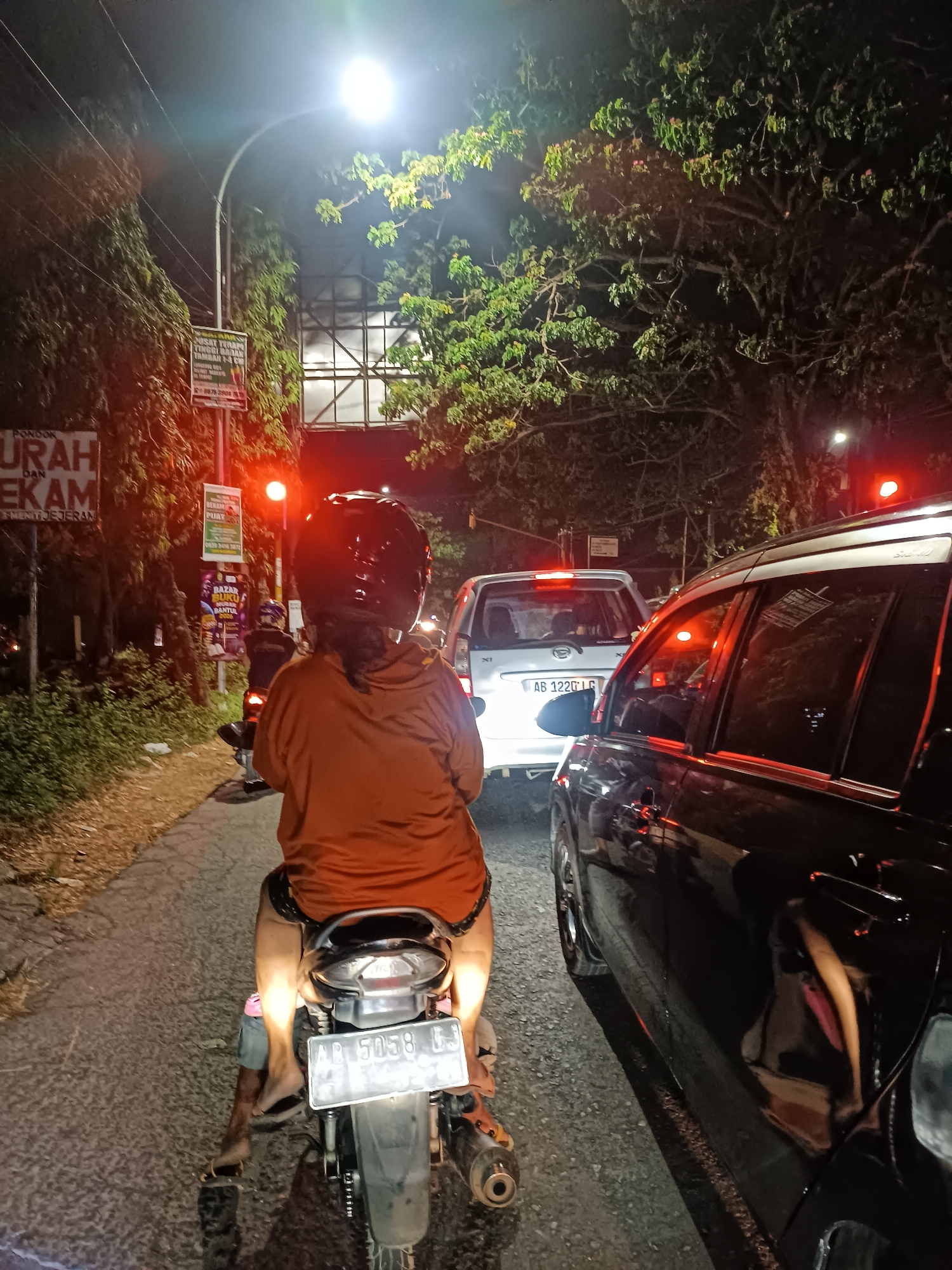 The image depicts a traffic jam at night on a road in Sewon, Indonesia. In the foreground, a person is riding a motorcycle, facing away from the camera. They are wearing an orange hooded jacket and black shorts. The motorcycle has a license plate that reads "AB 5058 GJ". The road is paved and appears wet, with visible tire tracks. Trees line the left side of the road, and a sign with Indonesian text is partially visible. In the background, several cars are stopped in traffic, including a silver SUV with a license plate "AB 1220 LG". Traffic lights are red, indicating that traffic is stopped. Overhead streetlights and vehicle headlights illuminate the scene.