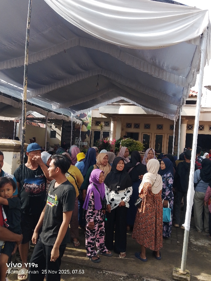 A group of people gathered beneath a large white canopy, appearing to be at an event or gathering. Most are wearing modest clothing and headscarves, suggesting a religious or cultural event. The building behind them has a neutral facade with symmetrical windows and columns. The atmosphere seems calm. The lower left corner of the image includes a time stamp.