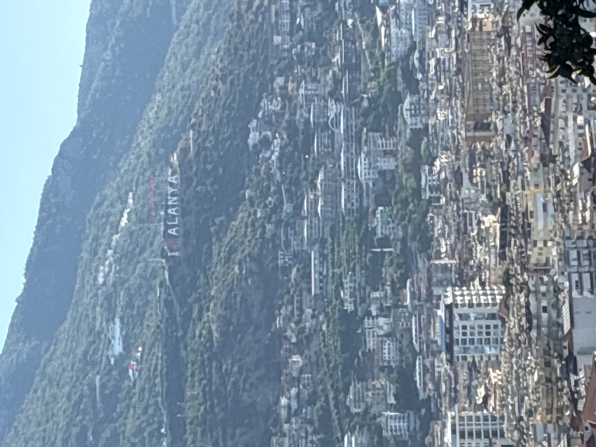 The image depicts a panoramic view of Alanya, Turkey, with a prominent "I LOVE ALANYA" sign on the hillside. The city is densely packed with buildings, predominantly in shades of white and beige, with some featuring balconies and multiple stories. The architecture suggests a Mediterranean style. The buildings are nestled on a steep, green hillside, indicating a town built on varied terrain. Above the cityscape, the hillside is covered with dense green vegetation, and the sky is clear blue. The overall scene conveys a sense of a well-established coastal city integrated with its natural landscape.