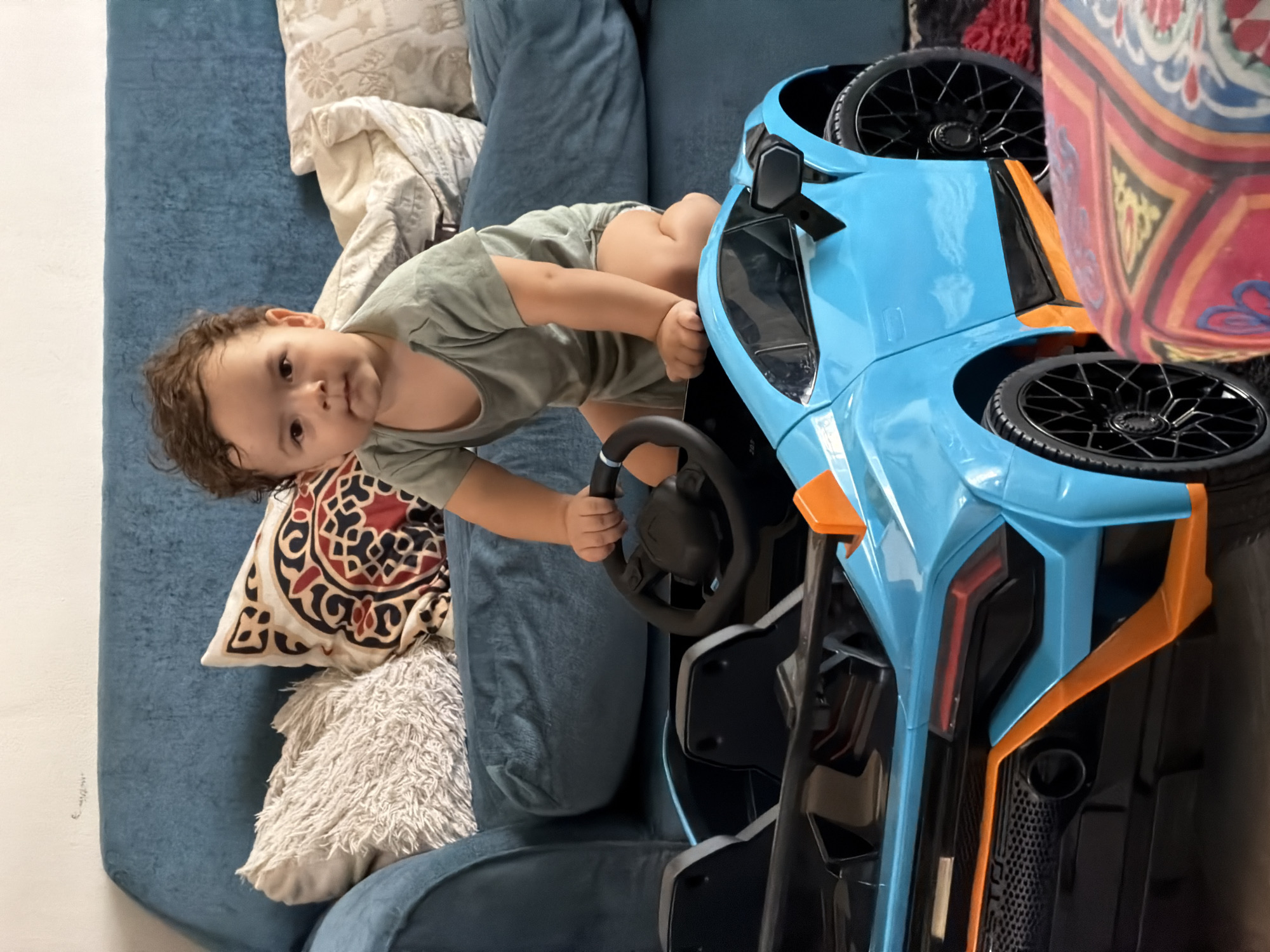 A young child with curly brown hair sits in a bright blue toy sports car. The car has orange accents and black wheels. The child is wearing a grey t-shirt and appears to be holding onto the steering wheel. The child is positioned on a plush blue sofa, which has several decorative pillows on it. One pillow has a red, black, and tan patterned design, and another is fluffy and white. The background suggests an indoor setting, likely a living room. This scene is not identifiable as being in Sharm El Sheikh, Egypt.