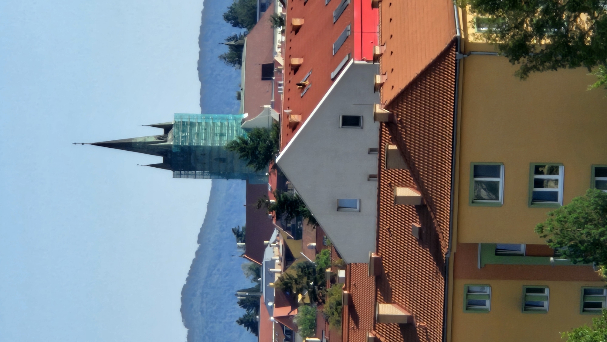This image captures a cityscape featuring a prominent church tower with a green scaffold, suggesting ongoing renovations. The tower's dark, pointed spire pierces the clear blue sky, with smaller spires flanking it. Surrounding the church are various buildings with red-tiled roofs, characteristic of many European cities. Some buildings appear to be residential, with visible windows and chimneys. In the foreground, a yellow building with green window frames is clearly visible, showcasing multiple stories and windows. The background is dominated by rolling hills covered in trees, creating a sense of depth. The overall scene depicts a relatively dense urban environment with a mix of architectural styles, set against a natural backdrop. The image appears to have been taken from an elevated position, offering a panoramic view of the city. The specific location in Prešov, Slovakia, is likely near the city center where historical buildings and residential areas coexist.