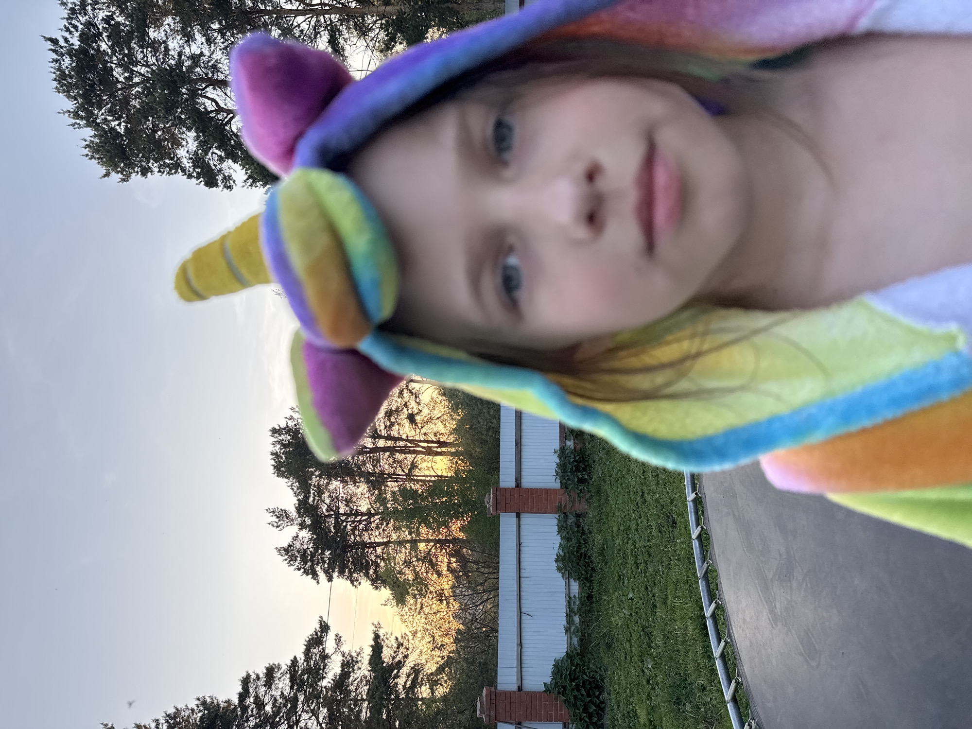 A young girl with fair skin and light eyes, wearing a colorful unicorn hooded robe, is positioned in the foreground of the image. The background features a yard with green grass and trees. A white fence with brick pillars is visible. The lighting suggests it is dusk or dawn. A trampoline is partially visible in the lower right corner of the image. It is not possible to verify the location.