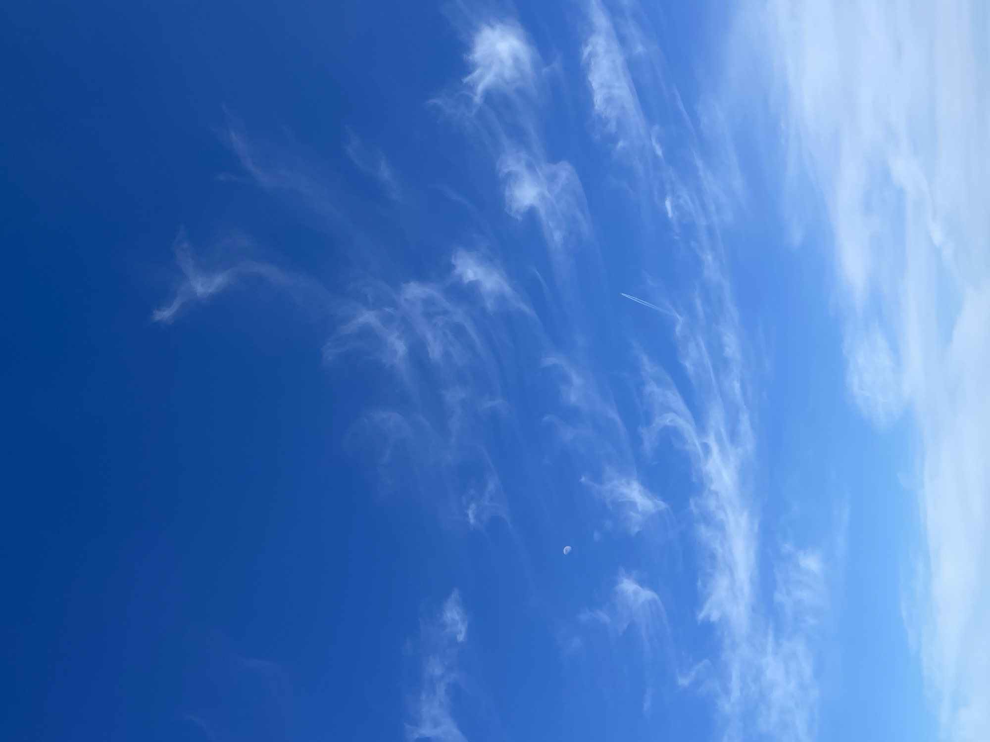 The image shows a daytime sky, predominantly blue, with streaks and patches of white clouds. The clouds are wispy and scattered. A faint contrail from a high-flying jet is visible. A small, pale object appears to be the moon. The location, Krasnoyarsk, Russia, is not directly discernable from the visual information within the image.