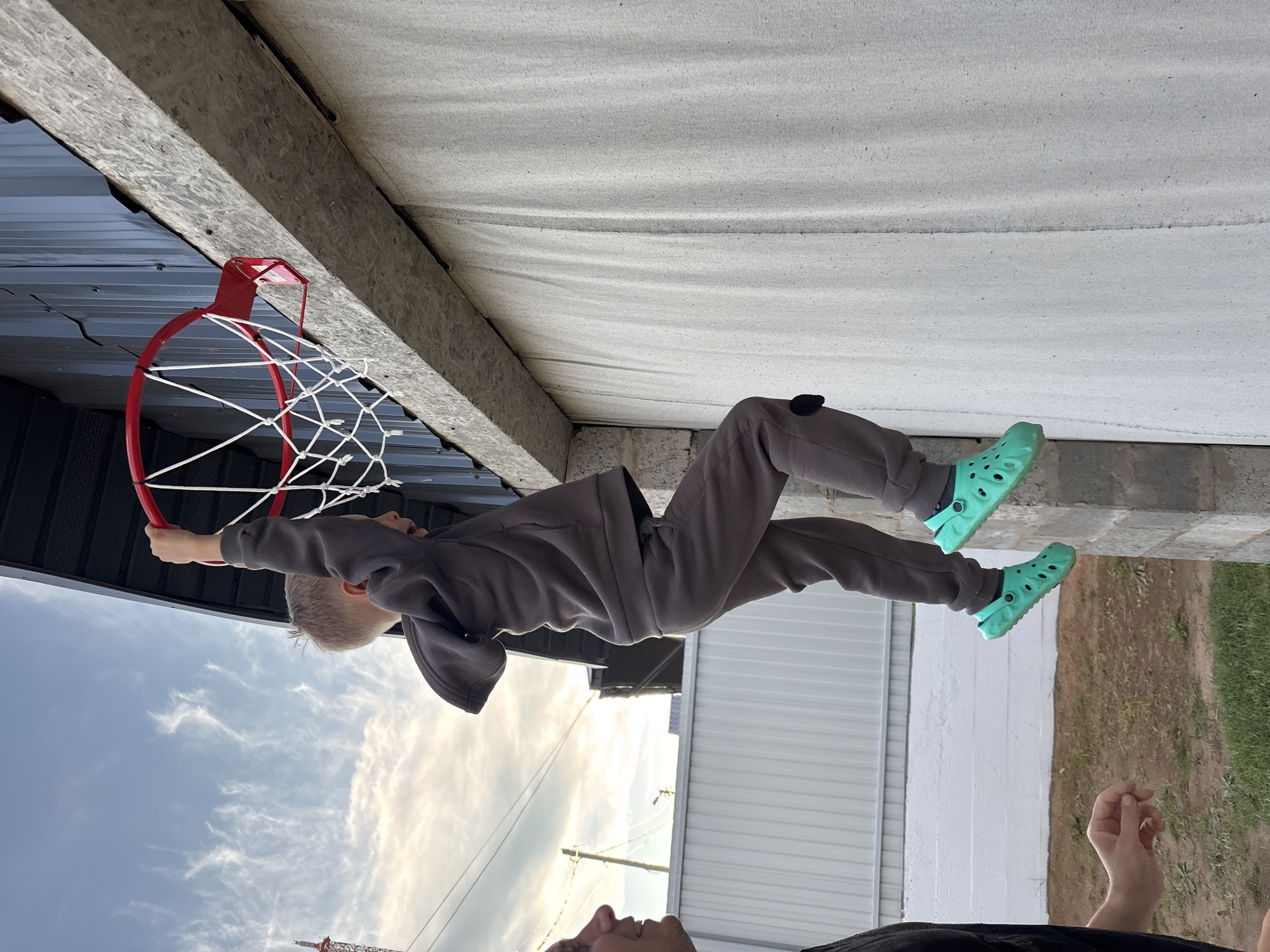 This image, taken in Chaikovsky, Russia, captures a dynamic outdoor scene featuring a young boy engaged in playful activity with an adult nearby. The central subject is a boy, dressed in a grey tracksuit and bright turquoise Crocs, who is hanging from a red basketball hoop with a white net. His arms are stretched upwards, grasping the hoop, and his mouth is open, suggesting excitement or effort.
The setting appears to be under an overhang or a structure with an exposed wooden beam and a light-colored, possibly fabric or textured, ceiling. In the background, there is a building with dark corrugated metal siding. The sky above is blue with scattered white clouds, indicating a clear or partly cloudy day, likely in the late afternoon or early evening given the soft lighting.
An adult's face and one hand are visible in the lower part of the frame, looking up at the boy, implying they are supervising or assisting him. The ground beneath is a mix of concrete and grass. In the far distance, faint power lines and a large red construction crane can be seen against the sky. The scene conveys a moment of active, joyful play. No visible text is present in the image.