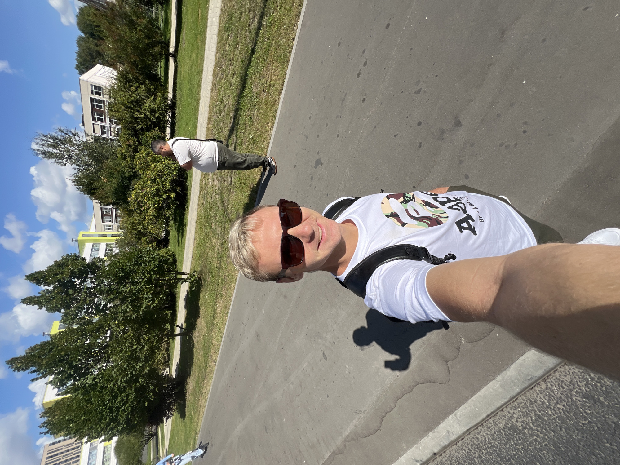 A man with blond hair and sunglasses is taking a selfie outdoors on a sunny day. He is wearing a white t-shirt with a camouflage pattern and the text "BAPE BY A BATHING APE", and dark pants. He has a backpack on. In the background, another man in a white t-shirt and dark pants is standing with his arms crossed, looking to the side. Behind him are green trees and modern buildings with yellow and white facades. The scene appears to be on a paved path, possibly a bike path or sidewalk, next to a grassy area. The sky is blue with some white clouds. The image seems to be taken in a park or urban area in Moskovskiy, Russia.