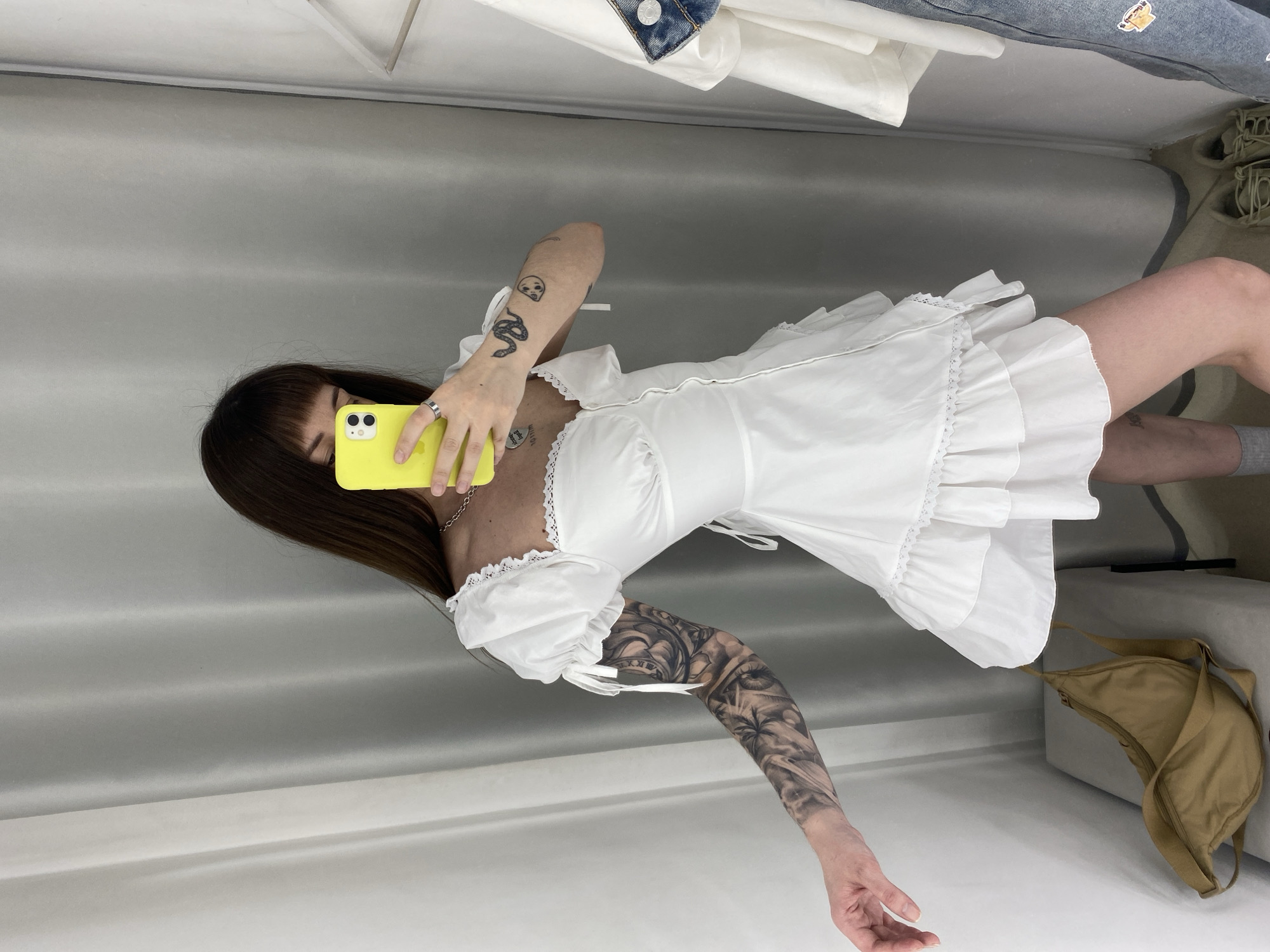 A person with long dark hair and bangs is captured in a mirror selfie, holding a bright yellow smartphone that partially obscures their face. They are wearing a white, short-sleeved mini-dress with ruffles, lace trim around the neckline, and a fitted bodice with lacing details. Light-colored socks are also visible.
Distinctive tattoos adorn both arms: the left arm features a detailed black and grey full sleeve, depicting intricate patterns including eyes and Roman numerals. The right forearm has several smaller black outline tattoos, including a snake, a stylized face, and another object. The person wears a silver ring on their left ring finger and a thin chain necklace from which the words "good girl" are faintly visible.
The setting appears to be an indoor space, likely a changing room or a well-lit room in Votkinsk, Russia. The background consists of light gray and white walls or curtains. In the upper right corner, a denim jacket with a partially visible "MYKON" tag and a small, cartoon-like patch hangs on a rail alongside other white clothing. A tan or khaki-colored shoulder bag rests on the floor or a bench in the lower right, next to a pair of light-colored sneakers. The lighting is bright and even, suggesting daytime.
