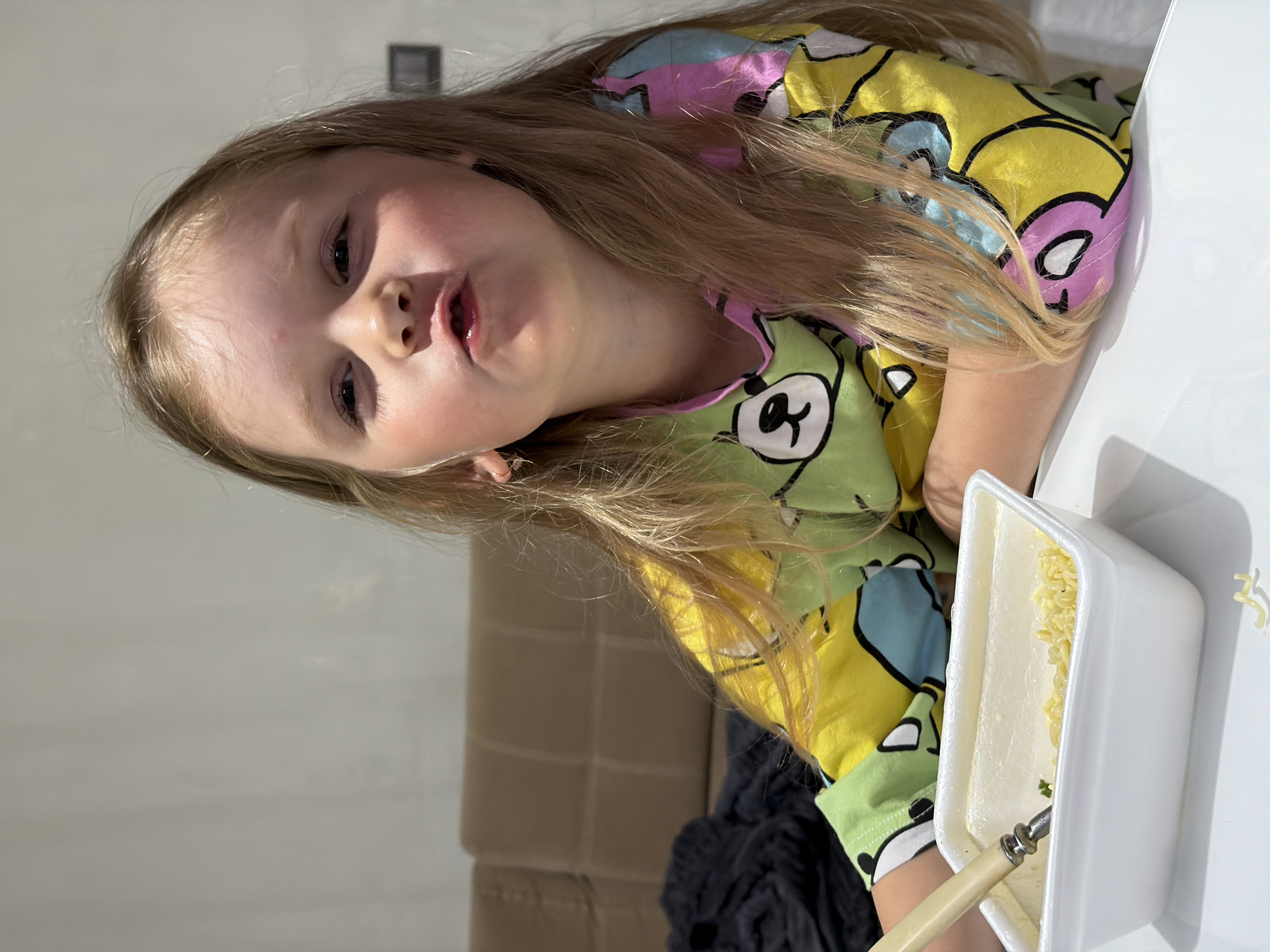 A young, fair-skinned child, appearing to be female with long, light blonde hair, is seated at a plain white table indoors. The child is wearing multi-colored long-sleeved pajamas patterned with cartoon bears in shades of green, yellow, light blue, and pink. Small gold hoop earrings are visible in her ears. Her lips are slightly pursed, and she looks directly at the camera with a neutral expression; a small red mark is present on her forehead.
In front of the child, a white rectangular foam or plastic container holds a portion of light-colored instant noodles with some green flecks, suggesting herbs or vegetables. A spoon with a beige handle and silver metal head rests in the container. Two detached cooked noodles are visible on the white table surface to the right of the container.
The background reveals a light brown or tan upholstered sofa to the left and directly behind the child. A dark, possibly black, textured fabric object or blanket is draped over an unseen item to the far left. The upper background consists of a plain, light gray wall, with a dark rectangular wall fixture (possibly a switch or outlet) visible on the right side. The visual evidence within the image provides no identifiable markers, signage, or architectural cues that would indicate its location as the city of Chaikovsky, Russia; it depicts a generic indoor residential setting.