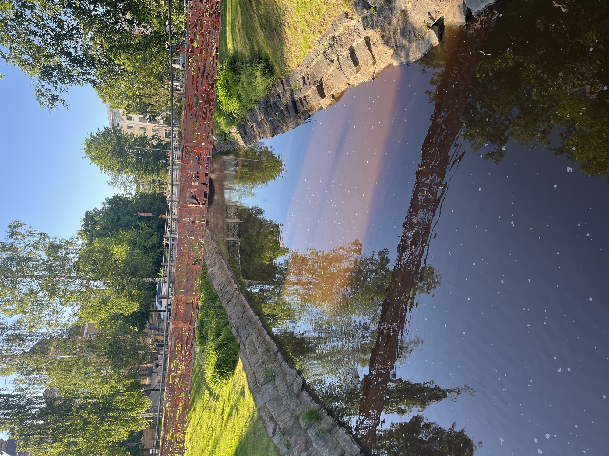 The image depicts a canal with a decorative red metal bridge spanning across it. The canal banks are lined with stone walls and grass. Trees and buildings are visible on either side of the canal, with a clear blue sky overhead. The water reflects the bridge, trees, and sky. The scene appears to be a public space within the city of Oulu, Finland, likely in a park or urban area.