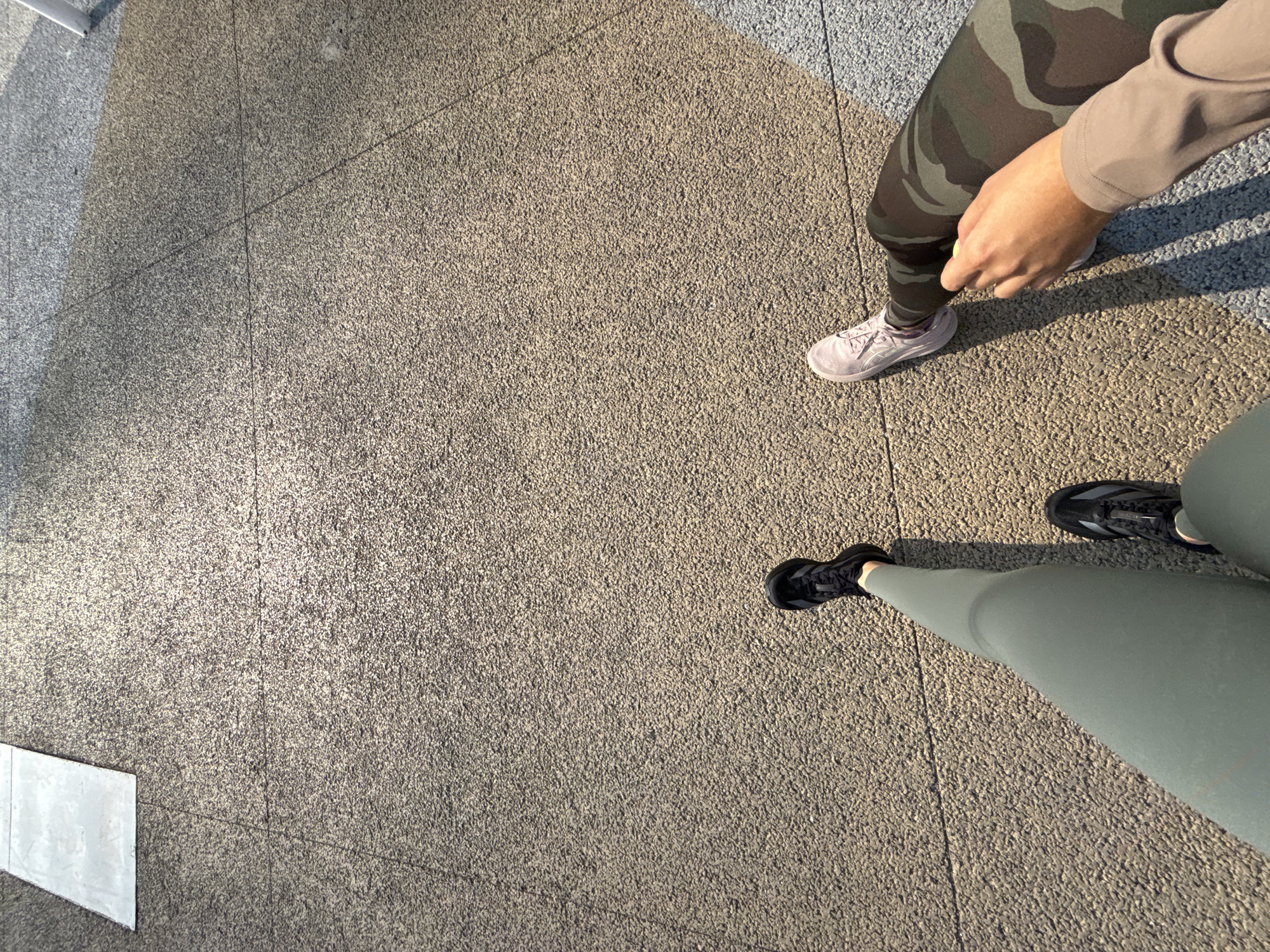 The image shows a view from above, focusing on the ground with two pairs of legs and feet. On the right side of the image is a person wearing camouflage leggings, a beige long-sleeved shirt, and pink sneakers. The second person is wearing green leggings and black athletic shoes. The setting appears to be an outdoor paved area, likely a sidewalk or public space. The environment includes concrete or stone paving, exhibiting a grainy texture. The image was probably taken in the city of Puebla, Mexico.