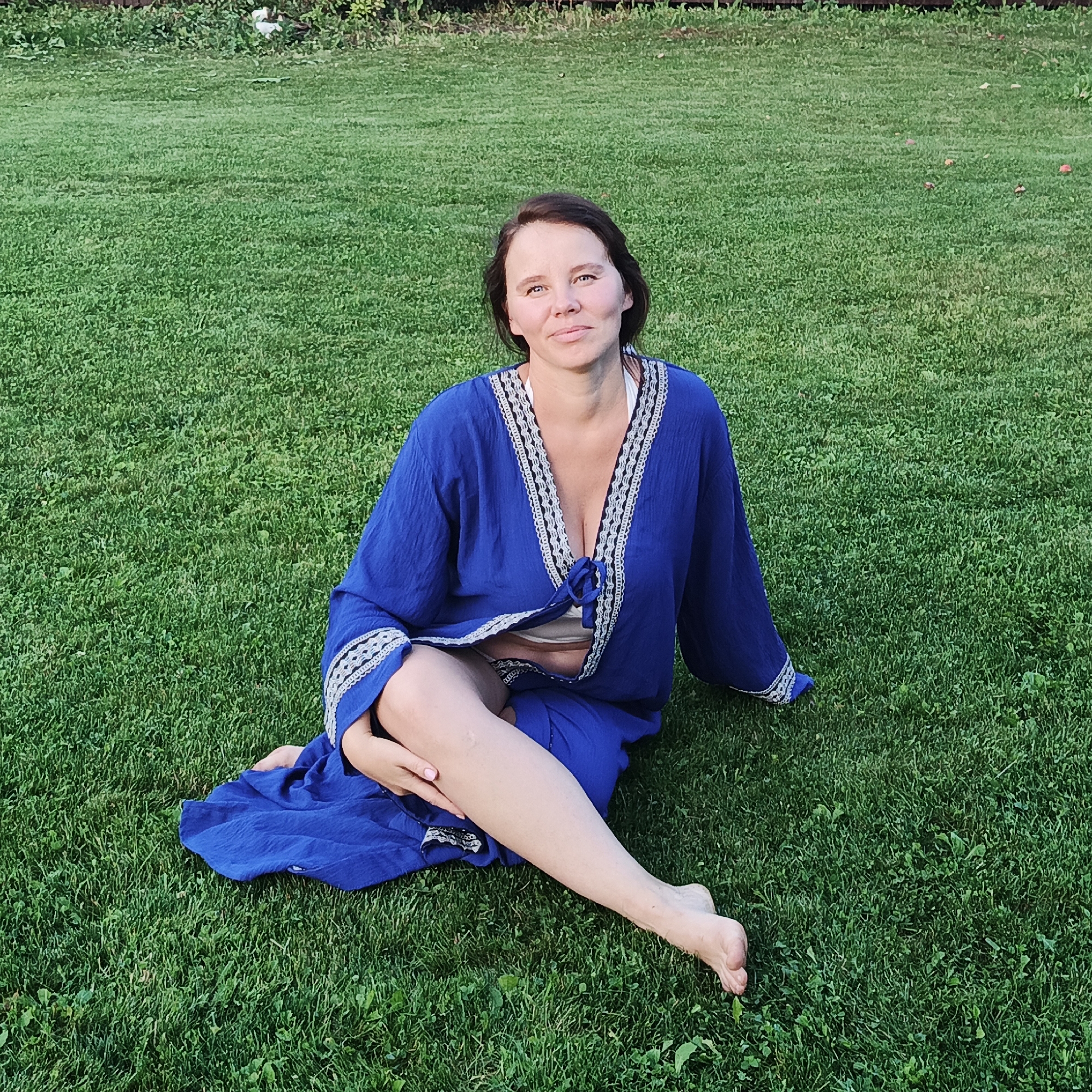 A woman with brown hair and light skin, wearing a blue robe with white and black trim over a white garment, sits on a green grassy lawn. Her legs are crossed, and her left leg is extended forward. The background consists of more green grass and a blurred line of vegetation. The lighting suggests it is daytime. The location is the city of Chaikovsky, Russia.