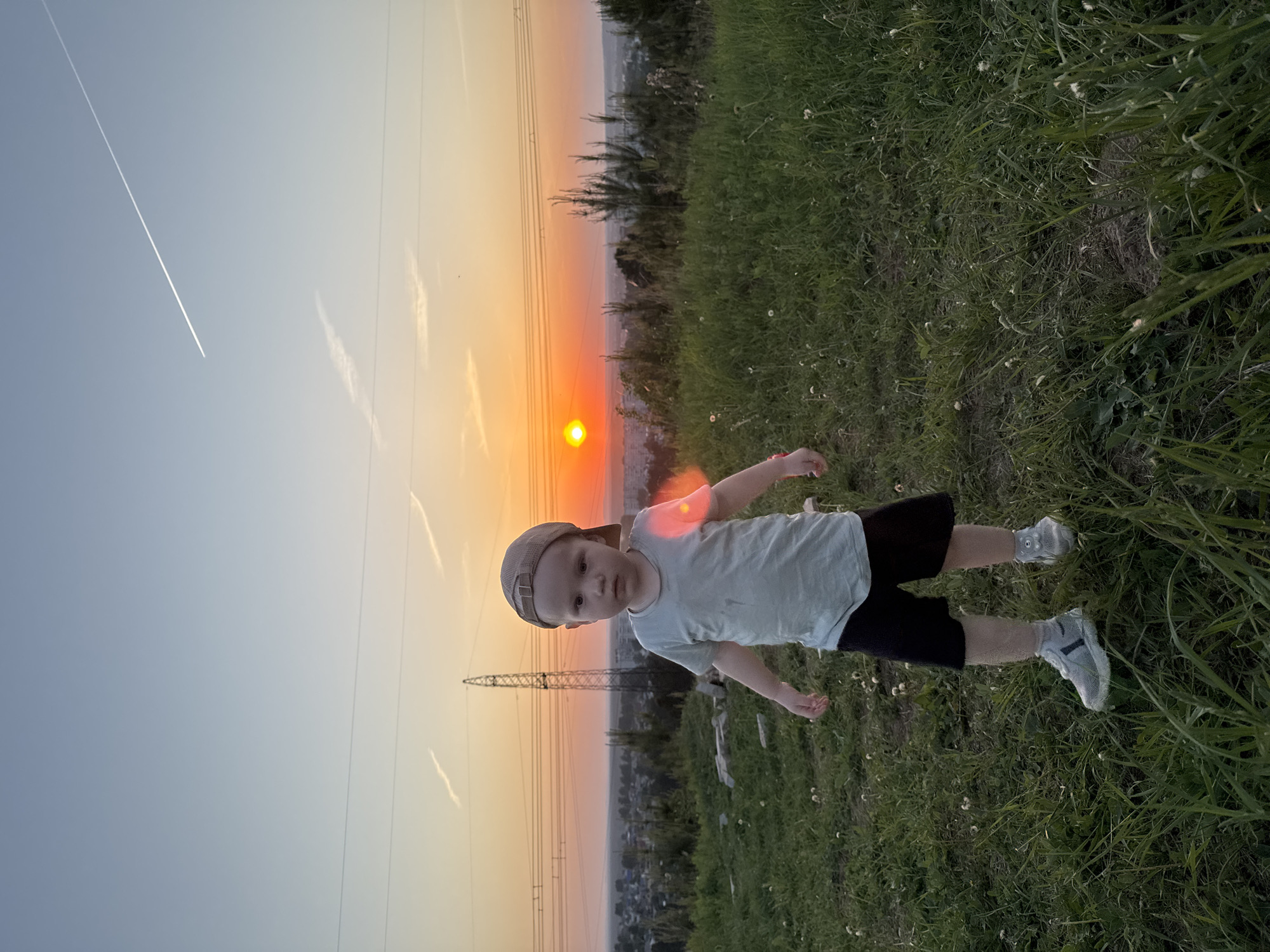 A young child stands in a field of tall grass at dusk. The child wears a baseball cap, t-shirt, and shorts, and is positioned in the foreground. Behind the child, a sunset casts a warm glow across the sky. A high-voltage power line tower is visible in the background, along with electrical lines stretching across the sky. In the distant background, the city of Chaikovsky, Russia can be seen, with buildings and vegetation visible in the twilight. A contrail from an aircraft is also visible in the sky.
