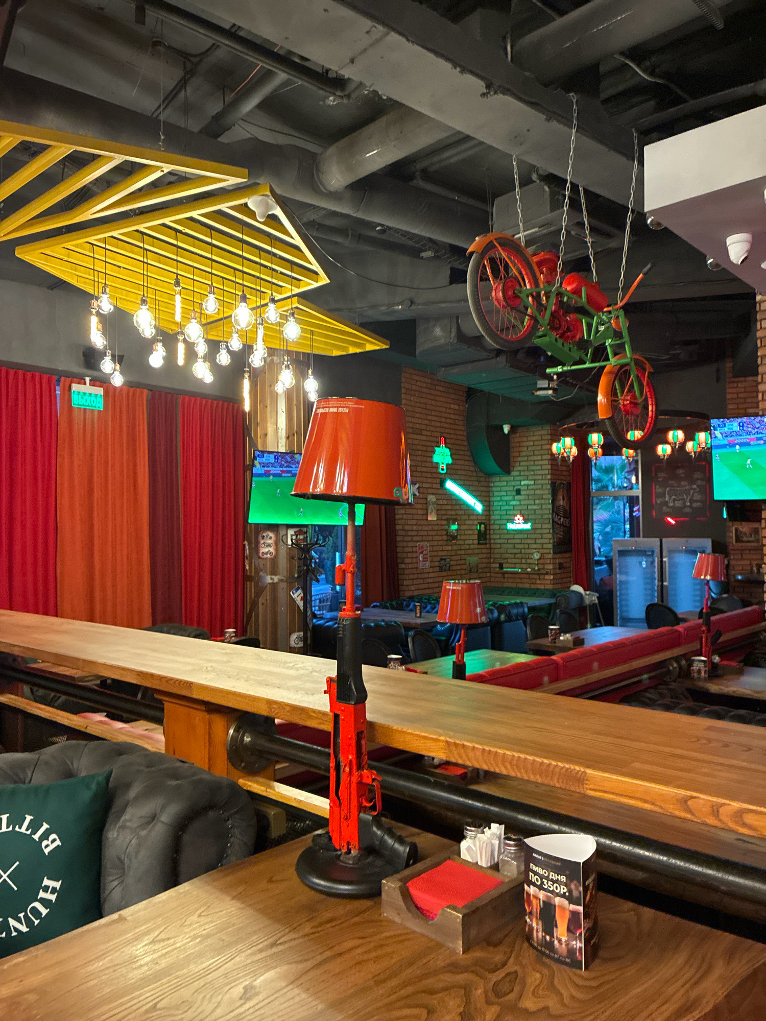 The image depicts the interior of a bar in Sochi, Russia. The scene is captured from a low angle, looking across a wooden counter towards the back of the establishment. The bar features a rustic and industrial aesthetic, with exposed brick walls, metal pipes, and wooden furniture.
Dominating the foreground is a long, light-colored wooden counter. On the counter, near the right edge, sits a decorative object resembling a red toy rifle, positioned in front of an orange lampshade. To the left of the rifle, a dark leather sofa is visible, adorned with a green cushion bearing white text. Further left, another section of the wooden counter extends.
The background reveals a variety of seating areas with red banquettes and dark leather chairs. Multiple televisions are mounted on the walls, displaying sports content. A vintage-style bicycle, painted orange and green, is suspended from the ceiling by chains. The ceiling itself is a dark, industrial expanse with visible ductwork and a prominent yellow geometric light fixture.
The lighting in the bar is a mix of exposed filament bulbs, pendant lights, and table lamps, contributing to a warm and inviting atmosphere. The overall impression is that of a lively bar or restaurant, possibly a sports bar given the presence of multiple televisions. The setting suggests a casual and social environment.