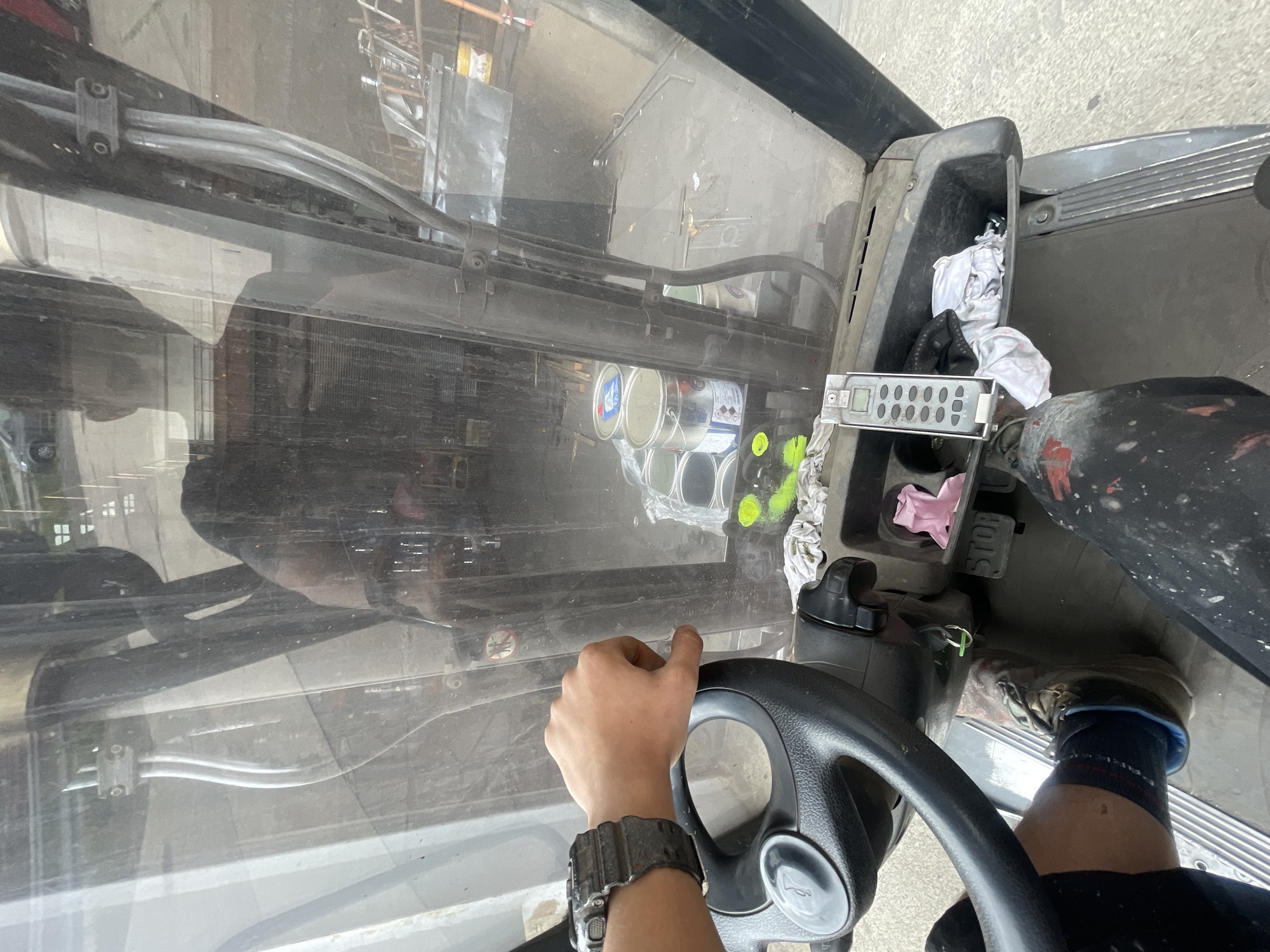 A first-person perspective shows a person's hand resting on the steering wheel of a forklift. The hand wears a black digital watch. The steering wheel is black and has a circular horn button in the center. The dashboard of the forklift is visible, with various items including a pink cloth, a remote control device with a keypad, and some crumpled white fabric. A pedal labeled "STOP" is visible near the floor. The person's legs are also visible, wearing dark pants splattered with white paint. The background shows a workshop or storage area with several metal cans, some appearing to contain paint. The scene is likely outdoors or in a well-lit indoor space, with reflections visible on the forklift's windshield, suggesting a somewhat overcast or dusty environment.