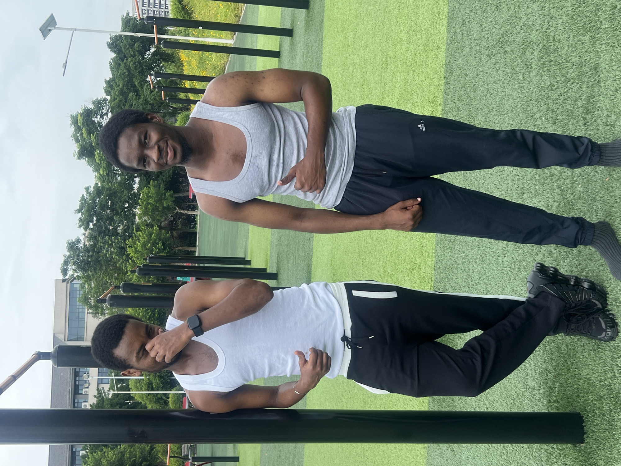 Two men are captured in a single frame; one standing and one leaning, appearing mirrored. The man standing is in a light gray tank top and dark Adidas pants. He is smiling and giving a thumbs-up. The man leaning is wearing a white tank top and black pants with white trim. He has a smartwatch on his left wrist and is covering his face with his left hand. The background reveals an outdoor workout area with a green artificial turf surface. There are black metal bars arranged in rows. A building and trees are visible in the distance, against an overcast sky. Based on visual cues and associated information, the scene is located in the city of Jiangjin, China.