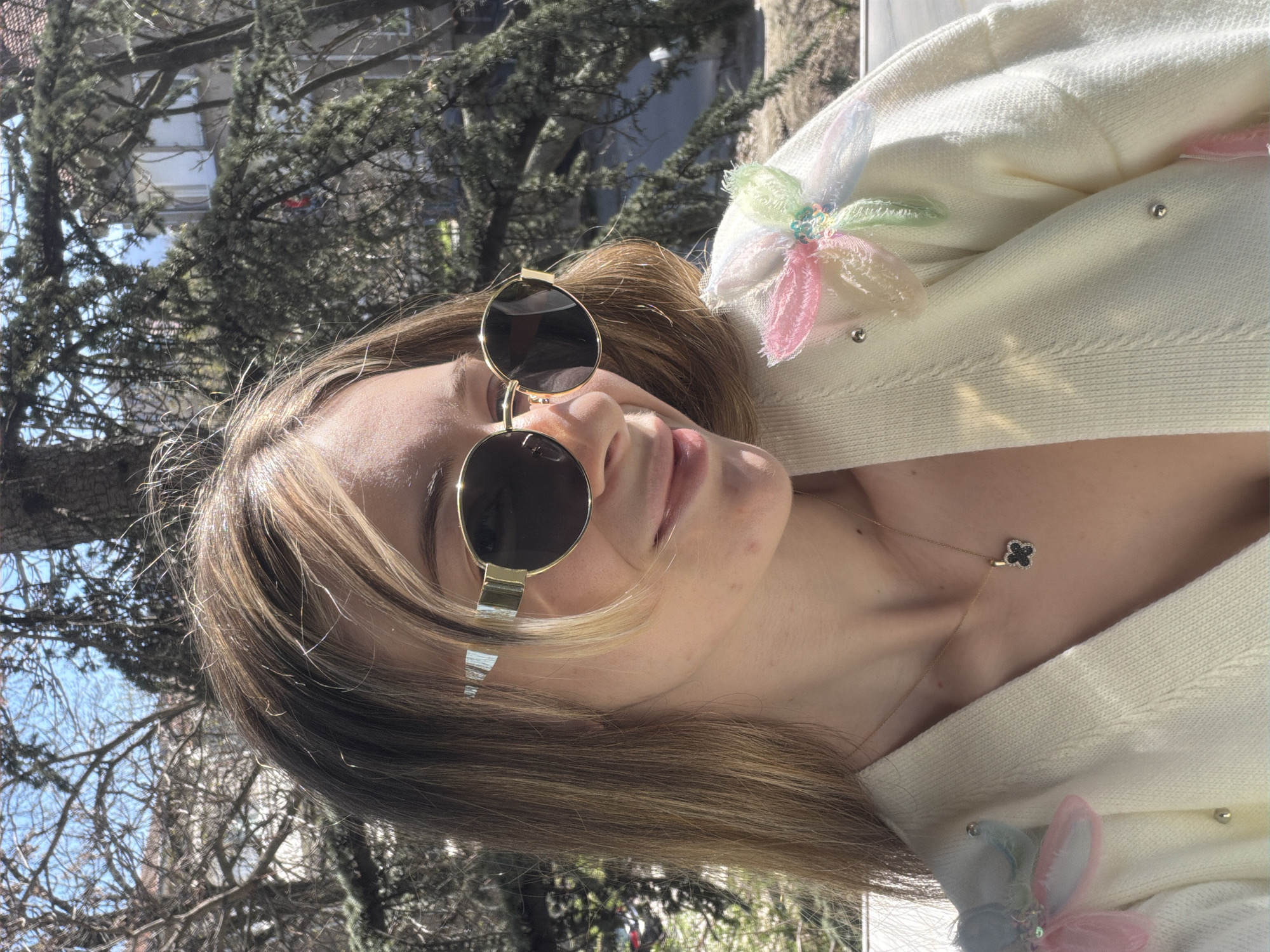 A young woman is posing in a sunlit outdoor setting, likely in Plovdiv, Bulgaria, based on the architectural details and vegetation visible in the background. She is wearing sunglasses and a cream-colored cardigan adorned with decorative flowers. A gold necklace with a black clover pendant is visible. The background includes a leafy tree and glimpses of buildings with windows under a blue sky.