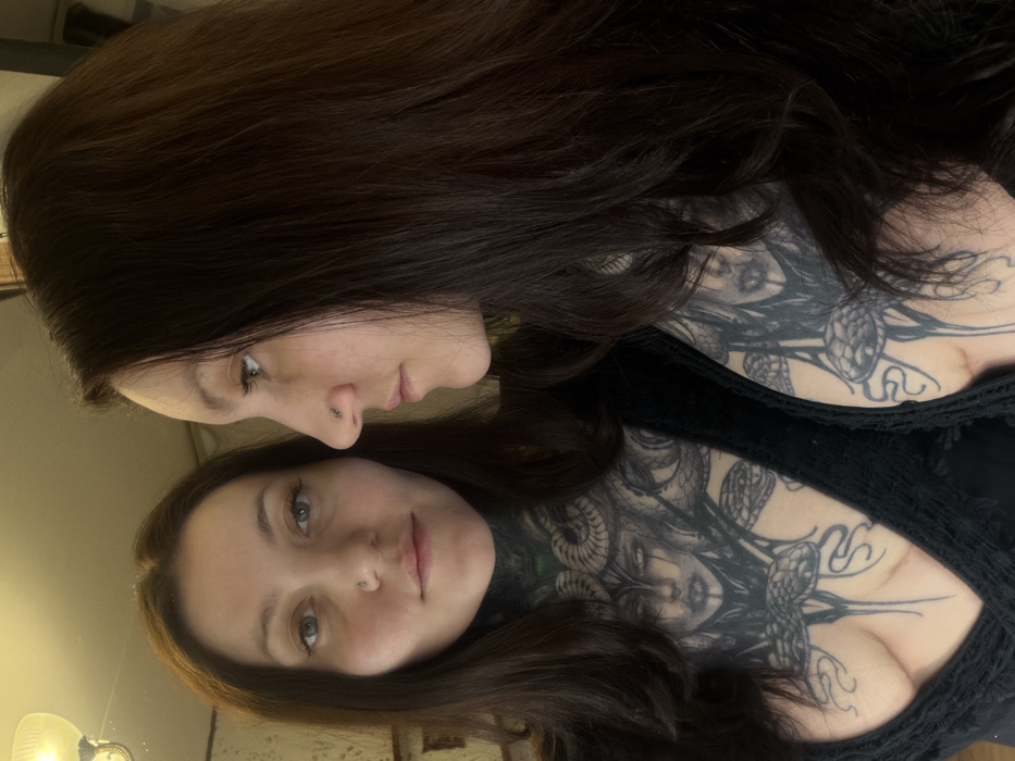 A woman with long, dark brown hair is captured in a selfie reflecting in a mirror. The top half of her body is visible, adorned with an elaborate tattoo of Medusa and snakes covering her chest and neck. She has fair skin, a nose piercing, and green eyes. Her reflection gazes directly at the camera, while she looks slightly to the side. She wears a dark, possibly lacy, top.