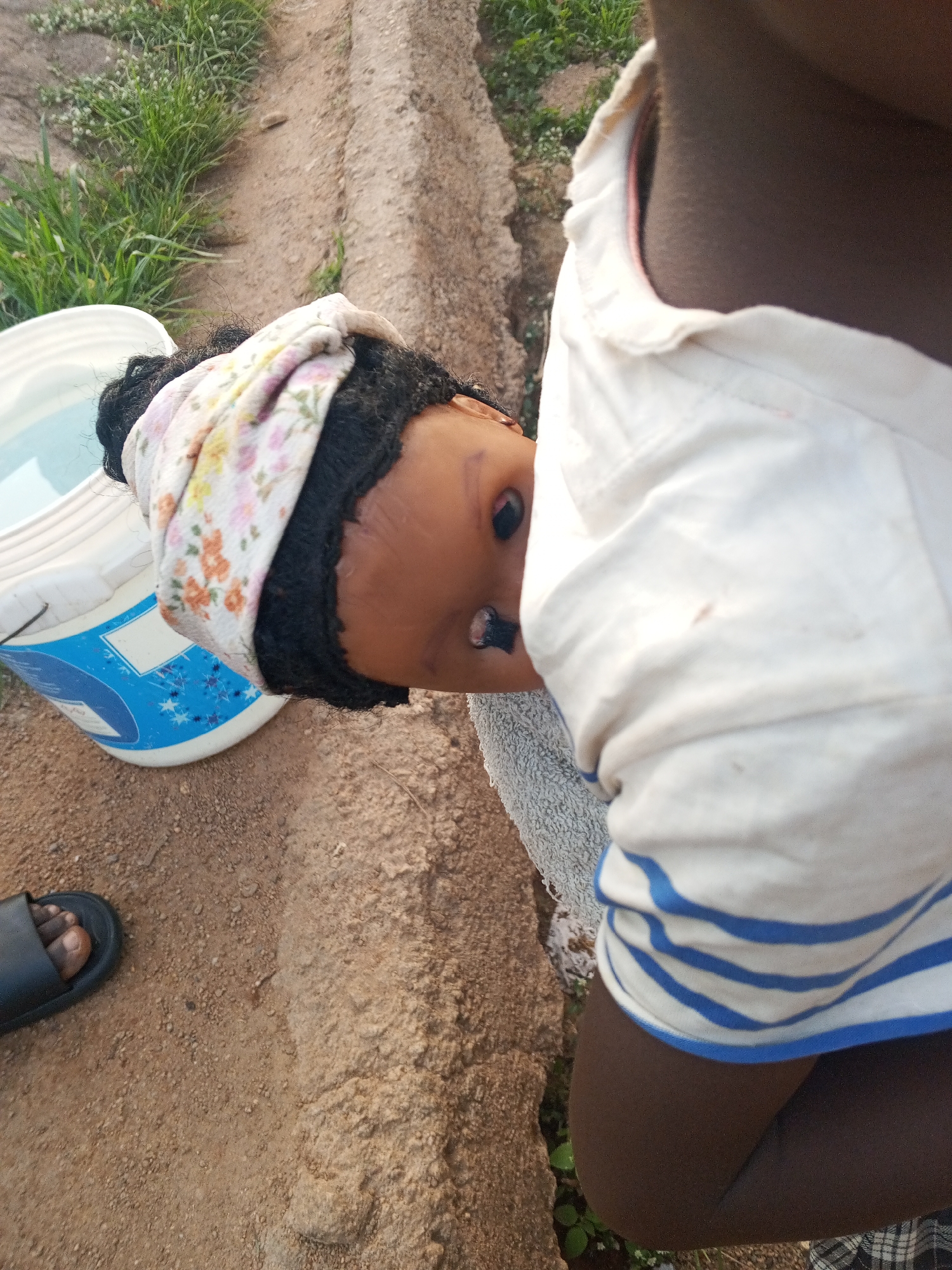 The image shows a dark-skinned child holding a doll. The doll has dark hair, is wearing a floral patterned headband, and is positioned so its face is visible. To the left of the doll is a white bucket with a blue design, and next to it, a foot wearing a black sandal is visible on the ground. The child is wearing a white shirt with blue stripes on the sleeves. The background consists of grass, dirt, and a small concrete wall or curb. The location is presumably Gwong, Nigeria.
