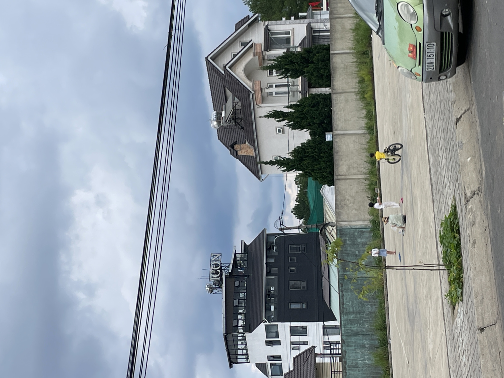 The image depicts an outdoor scene in Lâm Viên - Đà Lạt, Vietnam, under a cloudy sky. In the background, a modern, multi-story building with black and white sections features a prominent "ICON" sign on its rooftop. To its right is a large, light-colored house with a dark roof and architectural details. In the mid-ground, a concrete lot serves as a play area for several individuals. A child is riding a bicycle, and at least three other people, appearing to be adults or older children, are present, some engaged in activities on the ground. A small, young tree with green leaves stands in the foreground, rooted in a patch of greenery. A light green car is parked in the lower right portion of the frame, facing towards the left. A gray concrete wall runs across the mid-ground, separating the play area from the background buildings. Power lines are visible across the top of the image.
