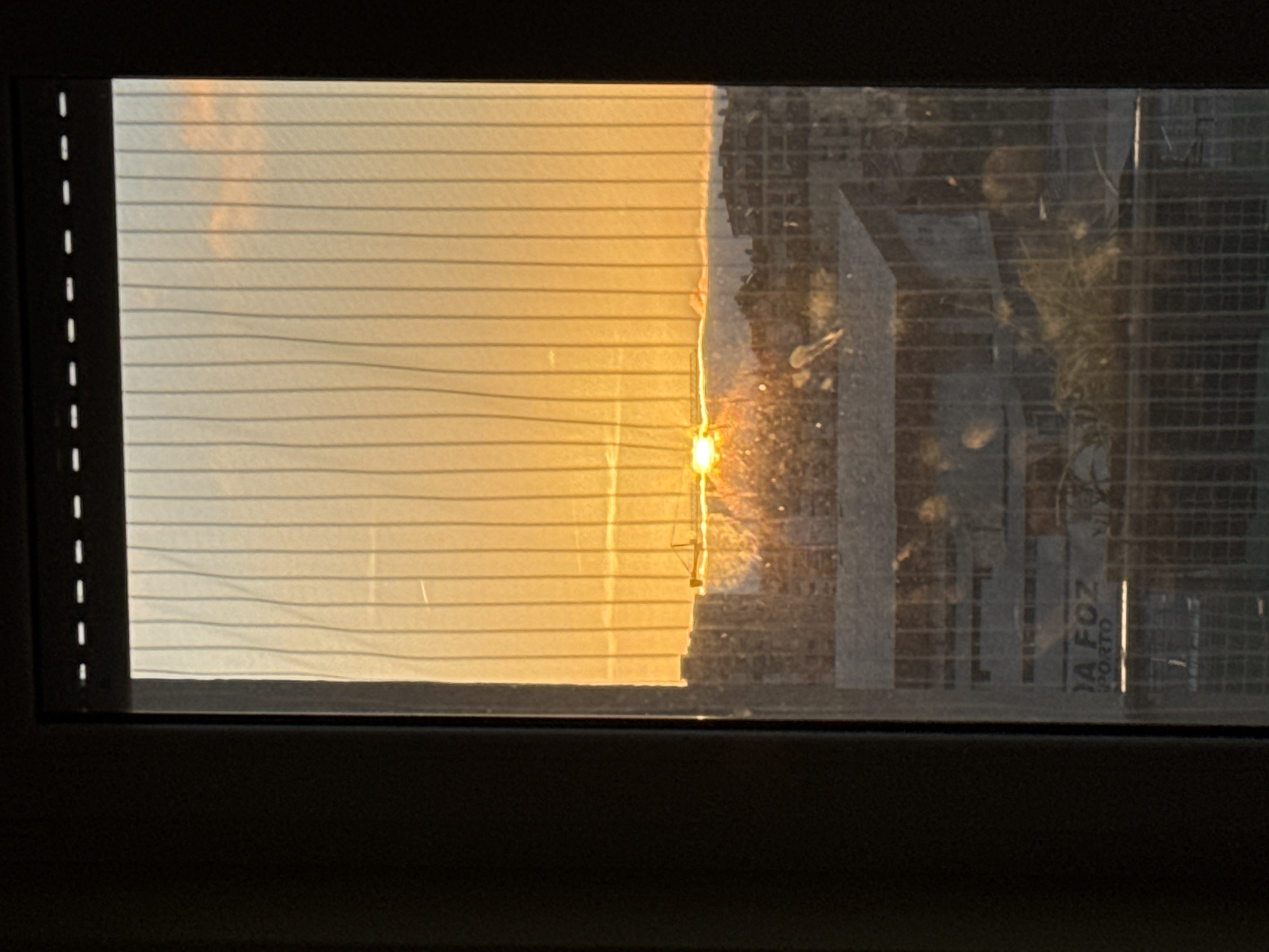The image depicts a sunset viewed through a window with horizontal blinds. Part of the blinds are pulled shut, and the sunset casts a warm, golden light on them. The rest of the view is a transparent window, where we can make out an urban landscape outside, with buildings and possibly foliage. There is writing on a poster on the right side of the image that says "DA FOZ PORTO". The image looks to be taken from inside a dark interior, framing the bright outdoor scene. Based on the text "DA FOZ" on the poster, it may be Figueira da Foz (or Porto).
