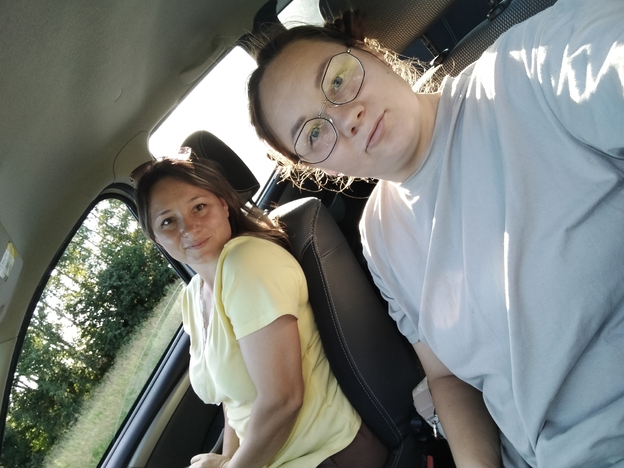 Two women are seated in the front seats of a car, likely in Russia, as indicated by the location context. The woman on the left, wearing a yellow t-shirt, has medium-length brown hair and is looking towards the camera with a slight smile. She is sitting in the passenger seat, with her right arm resting on the center console. The woman on the right, in a light gray t-shirt, is wearing glasses with a distinctive geometric frame and has her hair pulled back. She is looking directly at the camera, with a neutral expression. She is in the driver's seat, and her left arm is resting on her leg.

The setting appears to be daytime, with bright sunlight streaming into the car, creating strong highlights on the interior and on the woman on the right. The windows are open, and trees and a grassy field are visible outside, suggesting a rural or suburban location. The interior of the car is visible, including the seats, the ceiling, and part of the dashboard. There are no discernible emotions beyond slight smiles or neutral expressions. No text is visible in the image.
