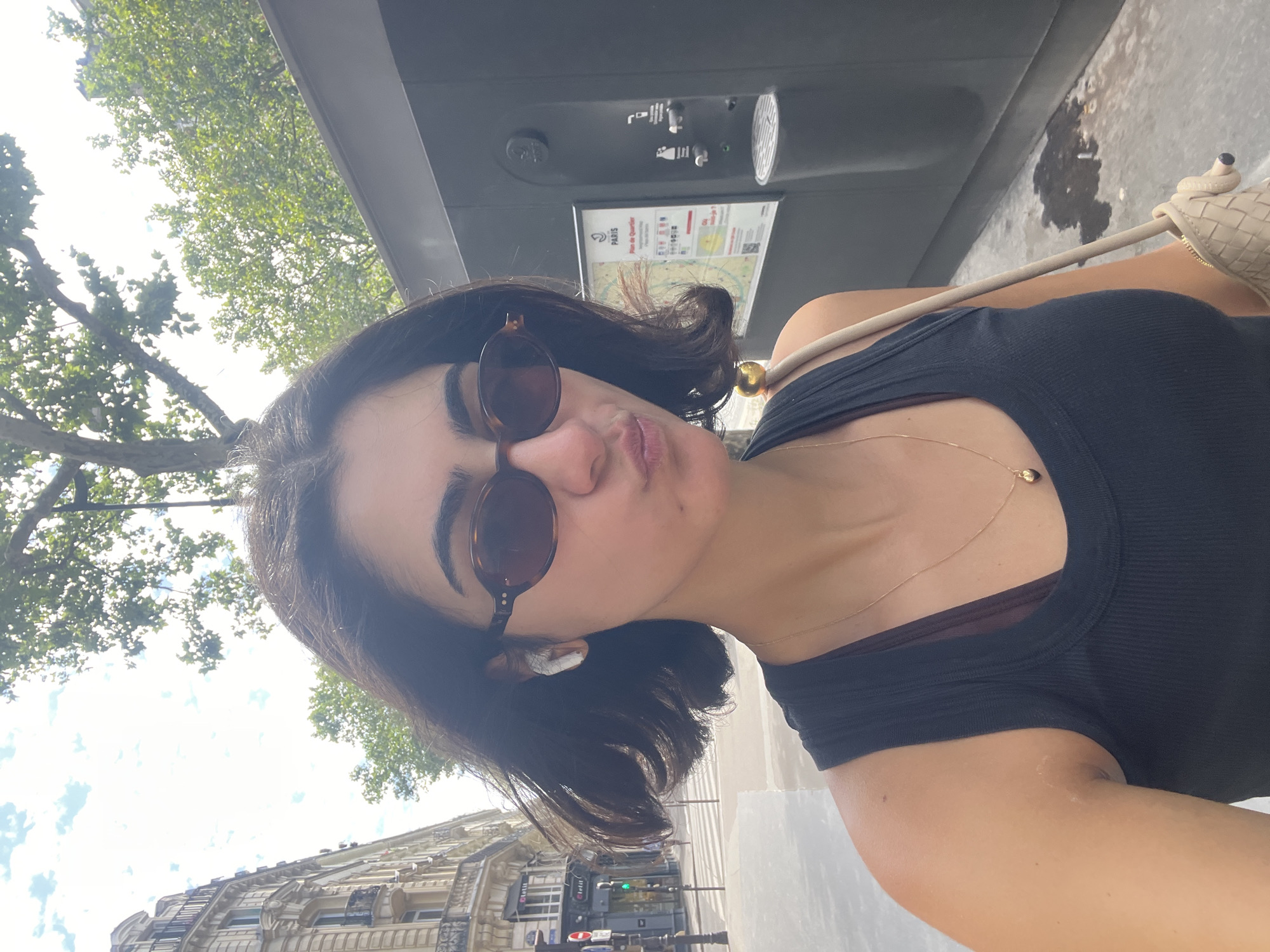In Paris, France, a young woman with dark hair styled in a bob is taking a selfie outdoors. She is wearing a black tank top, tortoiseshell sunglasses, and white earbuds. A delicate gold necklace with a heart-shaped pendant is visible. She is carrying a beige woven bag with a braided strap over her shoulder. The background features a classic Parisian building with balconies on the left and a modern, dark-colored structure with a water dispenser and a map of the neighborhood on the right. Trees with green foliage are present in the upper portion of the image. The ground appears to be a sidewalk.