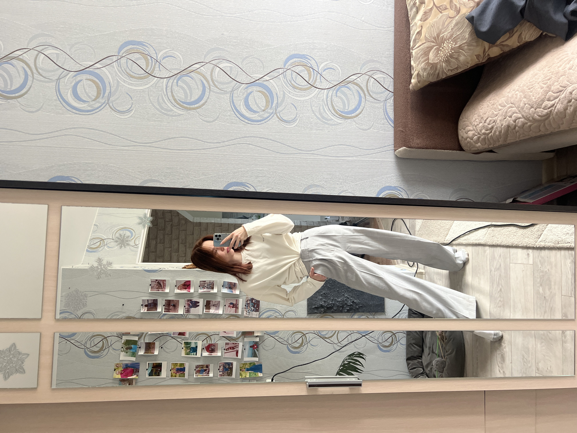 A young woman with reddish-brown hair is captured in a full-body mirror selfie, holding a smartphone with a light blue case. She wears a cream-colored long-sleeved top and light grey trousers, posing casually with one hand in her pocket. Her expression is neutral.
The setting is an indoor room, likely a home, featuring a large, multi-paneled mirror with a light-colored wooden or laminate frame. The mirror's surface is adorned with numerous small, square-format instant photos, depicting various people and possibly different scenes. Some panels of the mirror also show decorative snowflake motifs.
Reflected in the mirror, behind the woman, are glimpses of a textured wall, possibly brick or stone, and a dark, abstract artwork or panel. To her left (viewer's right in reflection), a green leafy plant is partially visible. The floor visible in the reflection features light wood-laminate flooring and a light-colored, shaggy rug with some wires on it.
Outside the mirror's reflection, the room's wall is covered in light-colored wallpaper (white or very light grey) with a repeating pattern of blue, grey, and brown swirling circles and wavy lines. To the right of the mirror, part of a bed or sofa is visible, covered in brown upholstery, with two pillows – one floral-patterned in beige tones and another light beige with an embossed design.
The lighting suggests daytime. The overall mood is relaxed and personal. No discernible text is visible on any objects within the image.