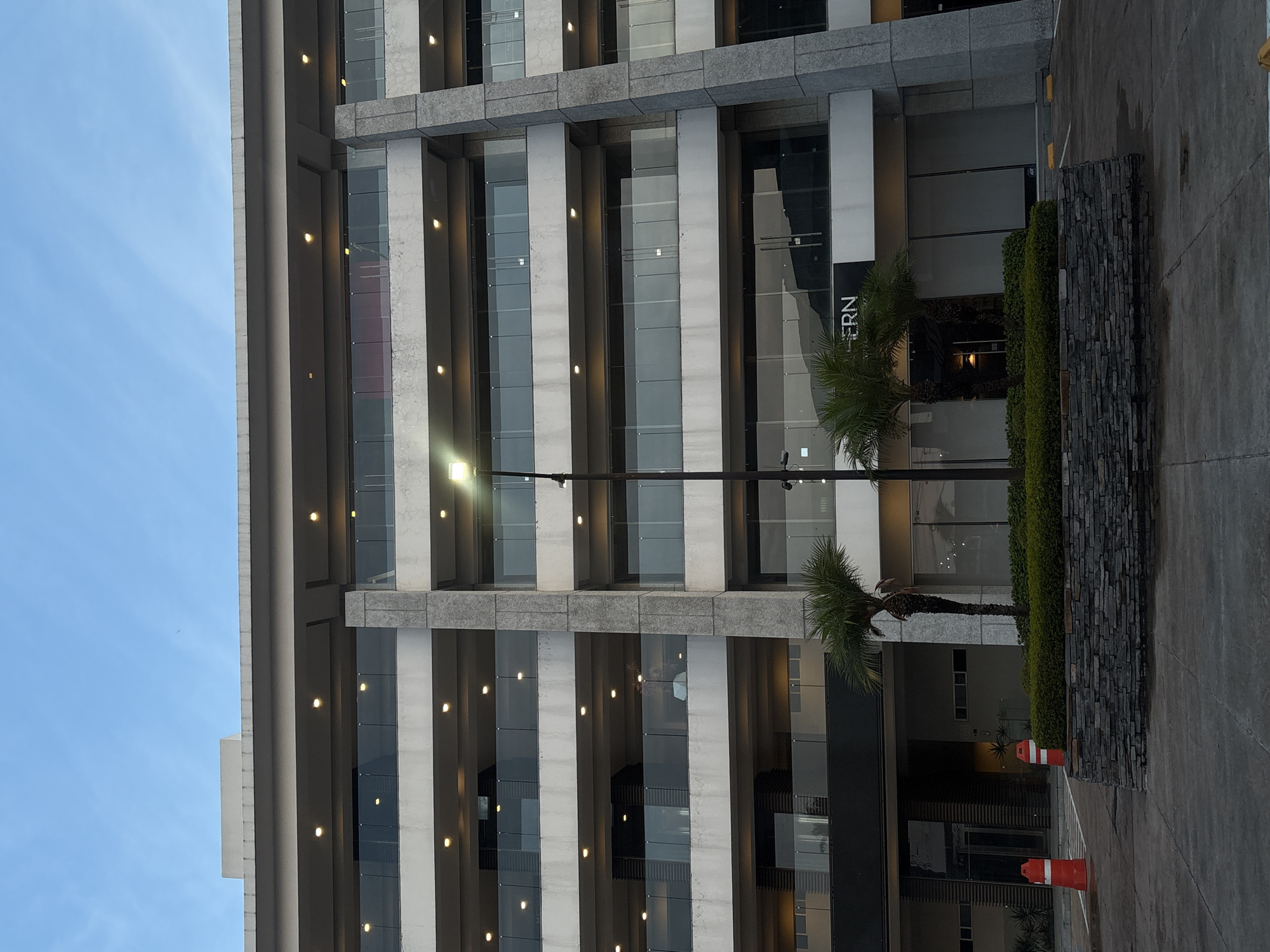 The image depicts the exterior of a multi-story building in San Bernardino Tlaxcalancingo, Mexico. The building features a modern architectural style with repeating vertical columns and large glass windows. There are rows of lights spaced evenly along the columns. In front of the building, there are two small palm trees and a low stone wall covered in greenery. Two orange traffic cones are positioned near the stone wall. The foreground is paved, and the sky is visible in the upper left corner, indicating daylight. A sign with the word "ERN" is partially visible on the building's facade.