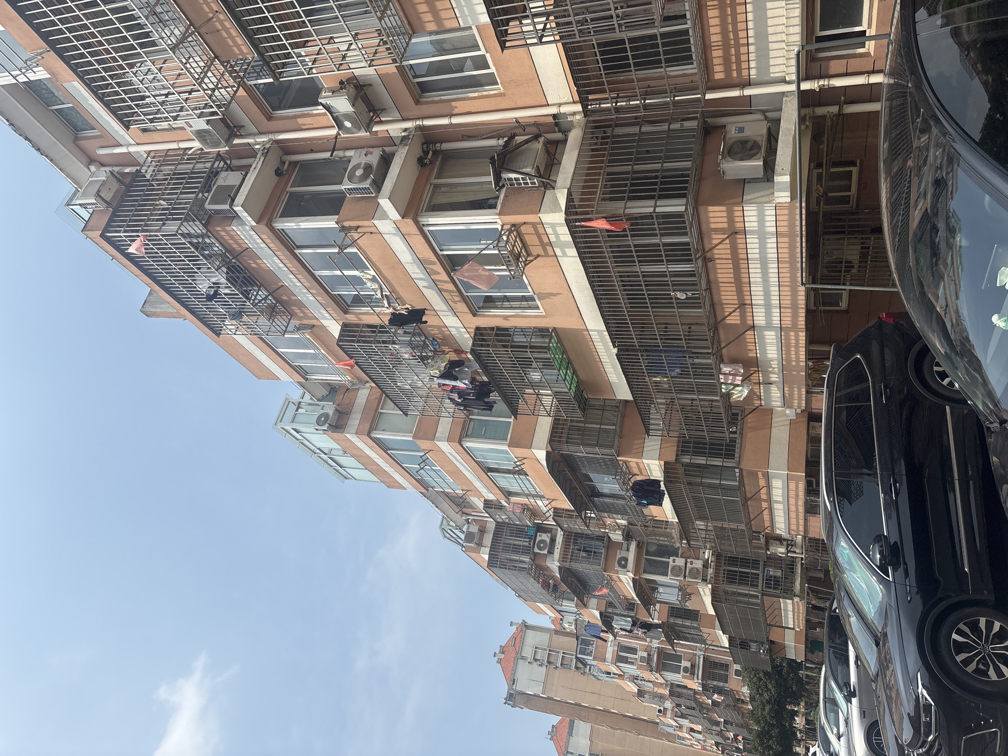 In the city of Suzhou Industrial Park, China, a multi-story apartment building with orange and white siding stands under a clear sky. The building features numerous windows and balconies, many of which are enclosed by metal bars. Laundry is hanging from clotheslines on several balconies. Air conditioning units are visible mounted outside on various floors. In the foreground, several cars are parked in a row, including a black SUV. The scene depicts a residential area with typical urban elements.