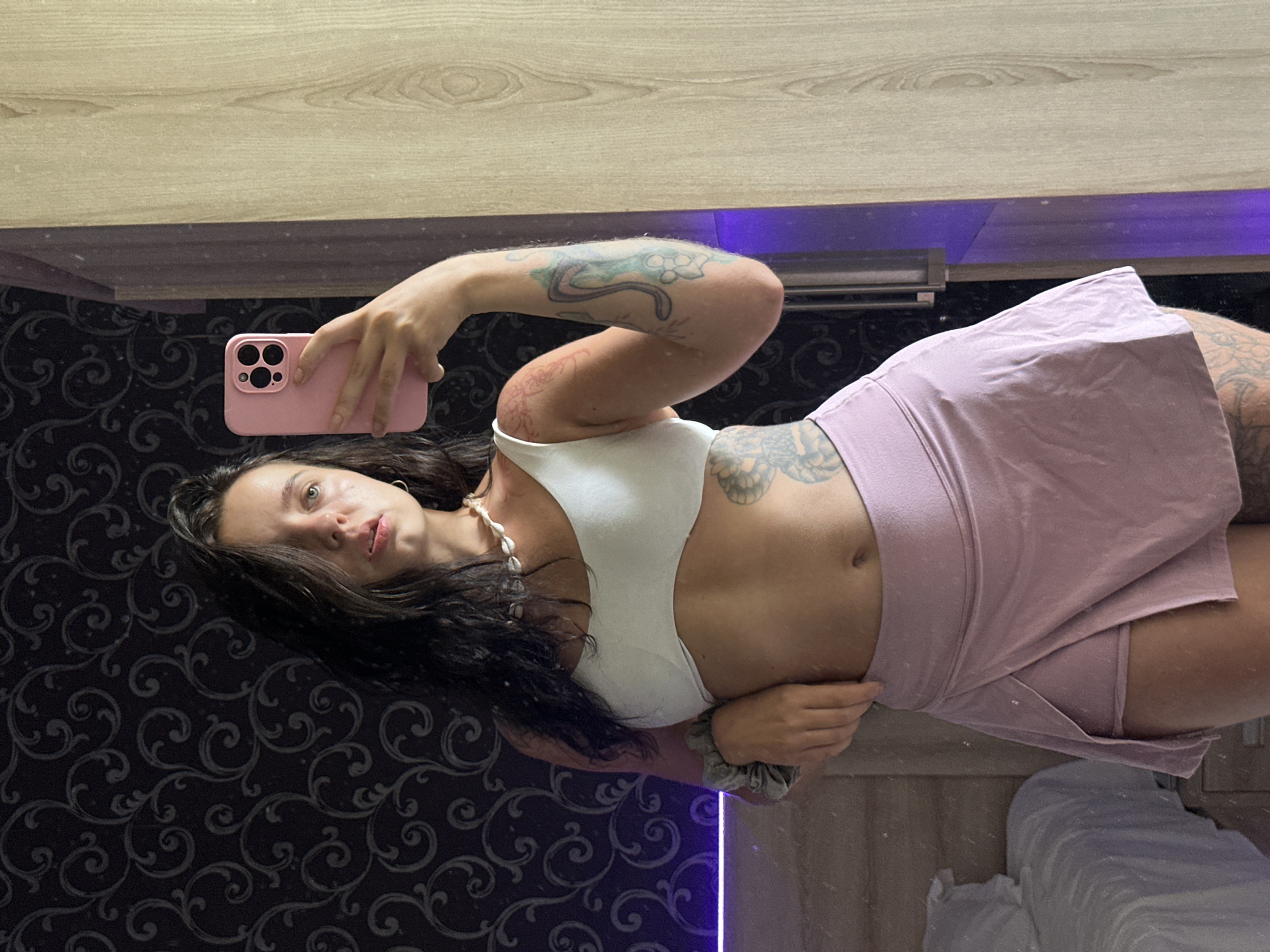 A young woman with dark hair and tattoos stands in front of a mirror, taking a selfie. She is wearing a white sports bra and a light pink skort. The background is a dark wallpaper with a swirling pattern. To the left, a bed with white sheets is visible, and to the right, a wooden closet door reflects the scene. A pink phone is held in her right hand, and her left hand is resting on her hip.