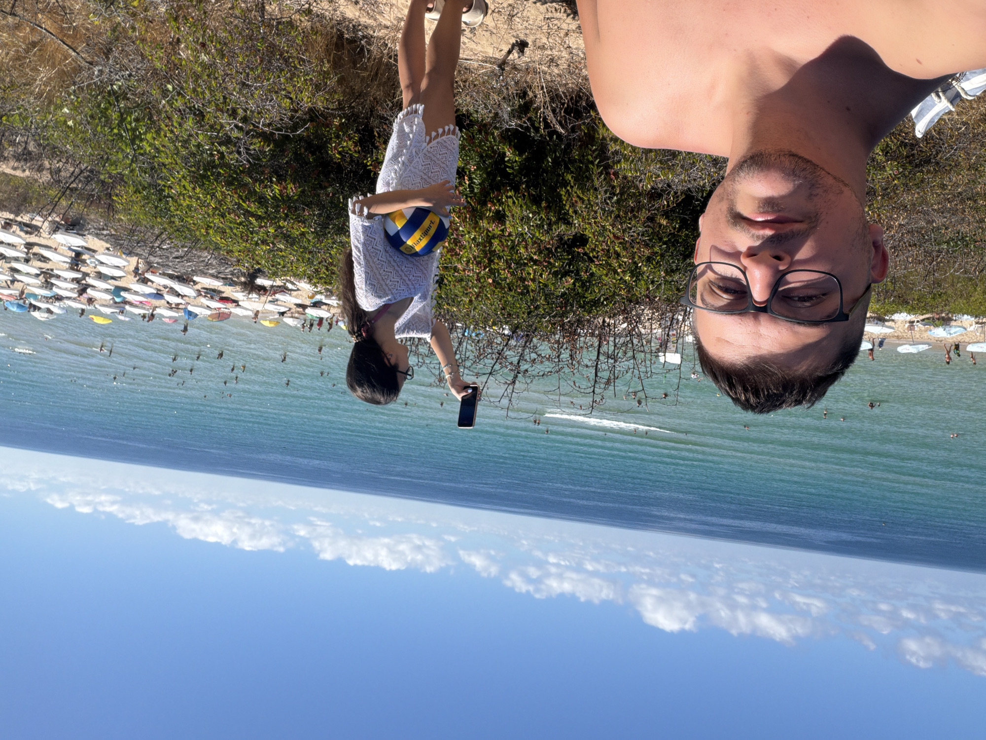 In the foreground, a man with short dark hair and a goatee, wearing glasses, smiles at the camera in a selfie. Behind him, a woman with dark hair and glasses holds a volleyball and a smartphone, appearing to take a photo. She is wearing a white cover-up. They are positioned on a sandy embankment covered with dry vegetation, overlooking a crowded beach. The beach is lined with numerous white and colored umbrellas. Beyond the beach, the turquoise sea is dotted with swimmers. The sky is a clear blue with scattered white clouds. The scene is located in Primorsko, Bulgaria, on a sunny day.