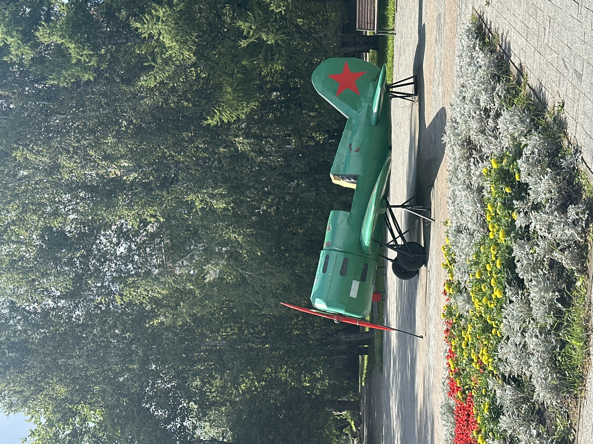 In an outdoor park setting, a weathered, bright green, single-engine propeller airplane is displayed on a paved walkway. The airplane features a prominent red propeller and a red star insignia on its tail fin. It is elevated on black metal stands. In the foreground, a well-maintained flower bed is visible, showcasing a vibrant arrangement of red, yellow, and silver-foliage plants. Behind the airplane, a row of mature green trees forms a lush backdrop, with some sunlight filtering through their leaves. A wooden park bench is situated to the right of the airplane, further into the park. The overall scene depicts a public space with a historical aviation exhibit and decorative landscaping.