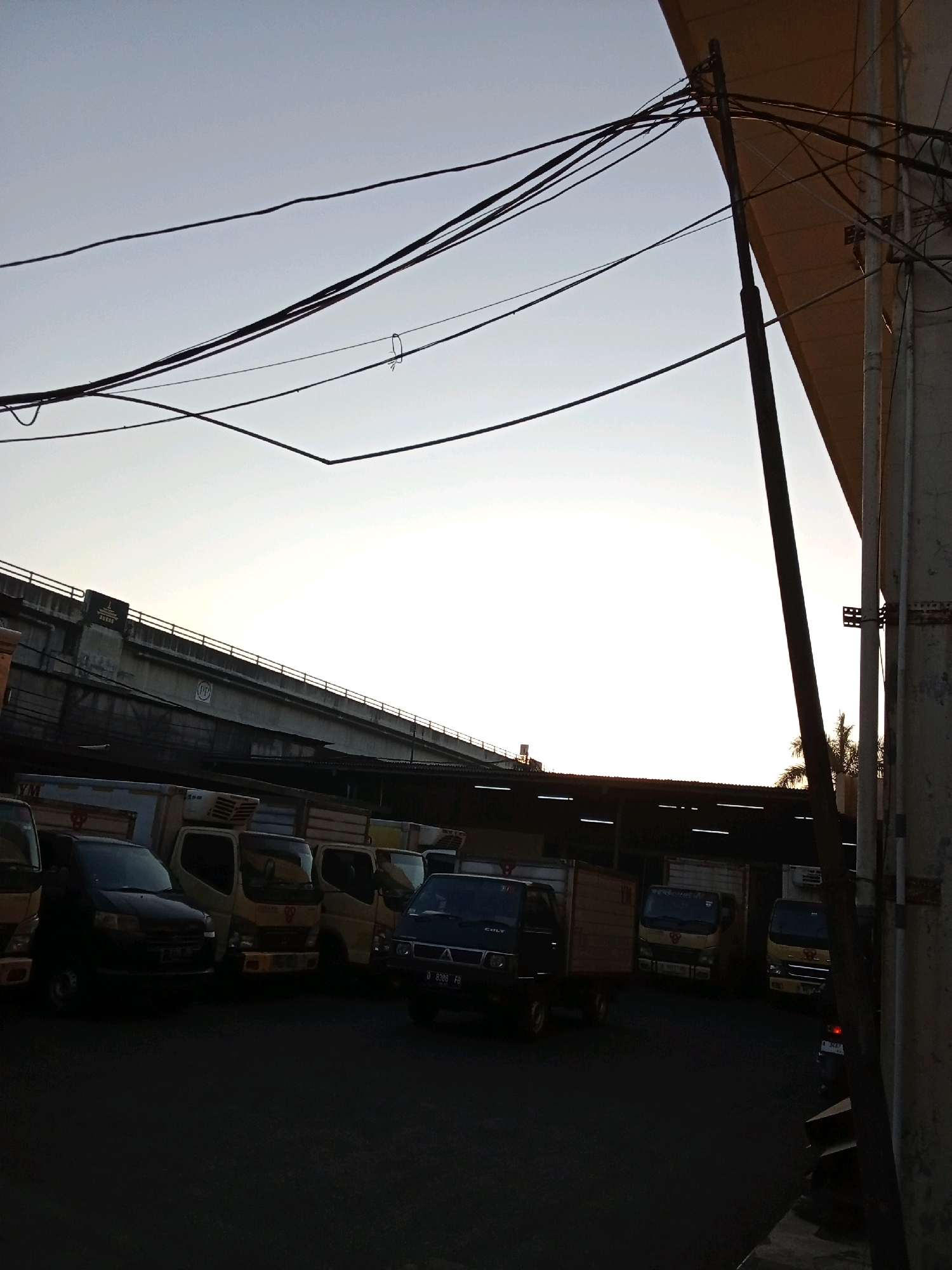 This image depicts an outdoor scene with several trucks parked in what appears to be a depot or a large parking area. The sky is bright and hazy, suggesting either early morning or late afternoon light. Overhead, a complex network of electrical wires crisscrosses the frame, attached to a dark pole.
In the background, a large concrete overpass or bridge dominates the upper portion of the image. Below the overpass, there is a row of buildings with corrugated metal roofs, and fluorescent lights are visible along the underside of these structures. The parked trucks are of various makes and models, mostly small to medium-sized commercial vehicles, some with enclosed cargo boxes and others with flatbeds. One black car is parked in the foreground, and a small motorcycle is partially visible on the right side of the frame.
The ground is paved and dark. The overall impression is one of a working area or logistics hub, possibly in an industrial or commercial district. The presence of the overpass and the type of vehicles suggest an urban setting.
The specific location in Bandung City, Indonesia, is not definitively identifiable from the visual elements alone. However, the overall context of parked commercial vehicles, overpasses, and urban infrastructure is consistent with many such areas within the city.