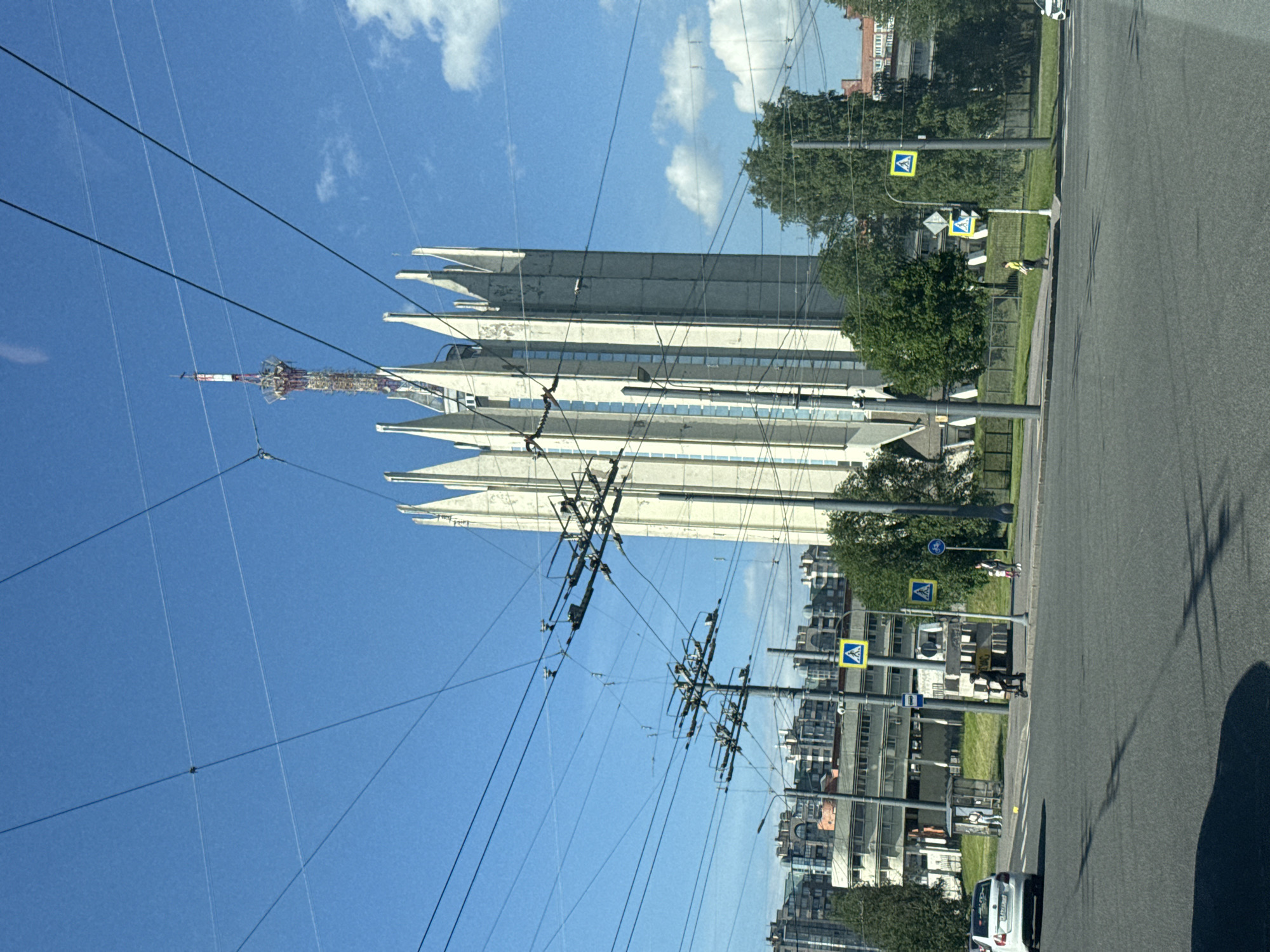 The image displays a sunlit urban street scene in Saint Petersburg, Russia, on a clear day with a bright blue sky and scattered white clouds. On the left side, a prominent modern building with a distinctive, pointed, or jagged facade dominates the view. This light-colored, multi-story structure is adjoined by a tall, lattice-style telecommunications tower, which rises above the building and is covered with antennae and equipment. Several faint aircraft contrails are visible high in the sky.
Numerous black overhead power lines or trolleybus wires crisscross the upper and middle portions of the frame, indicating active urban infrastructure. To the right of the wires and buildings, an asphalt road stretches into the distance. A white car is partially visible on the road in the lower right corner. A grassy median and sidewalk separate the road from the buildings, lined with green trees.
Along the street, several poles support yellow diamond-shaped pedestrian crossing signs, featuring a black silhouette of a walking person. One person wearing a bright yellow safety vest is visible near one of these pedestrian crossings. Another person in dark clothing can be seen further down the street. The scene captures a typical daytime moment in a bustling city environment.