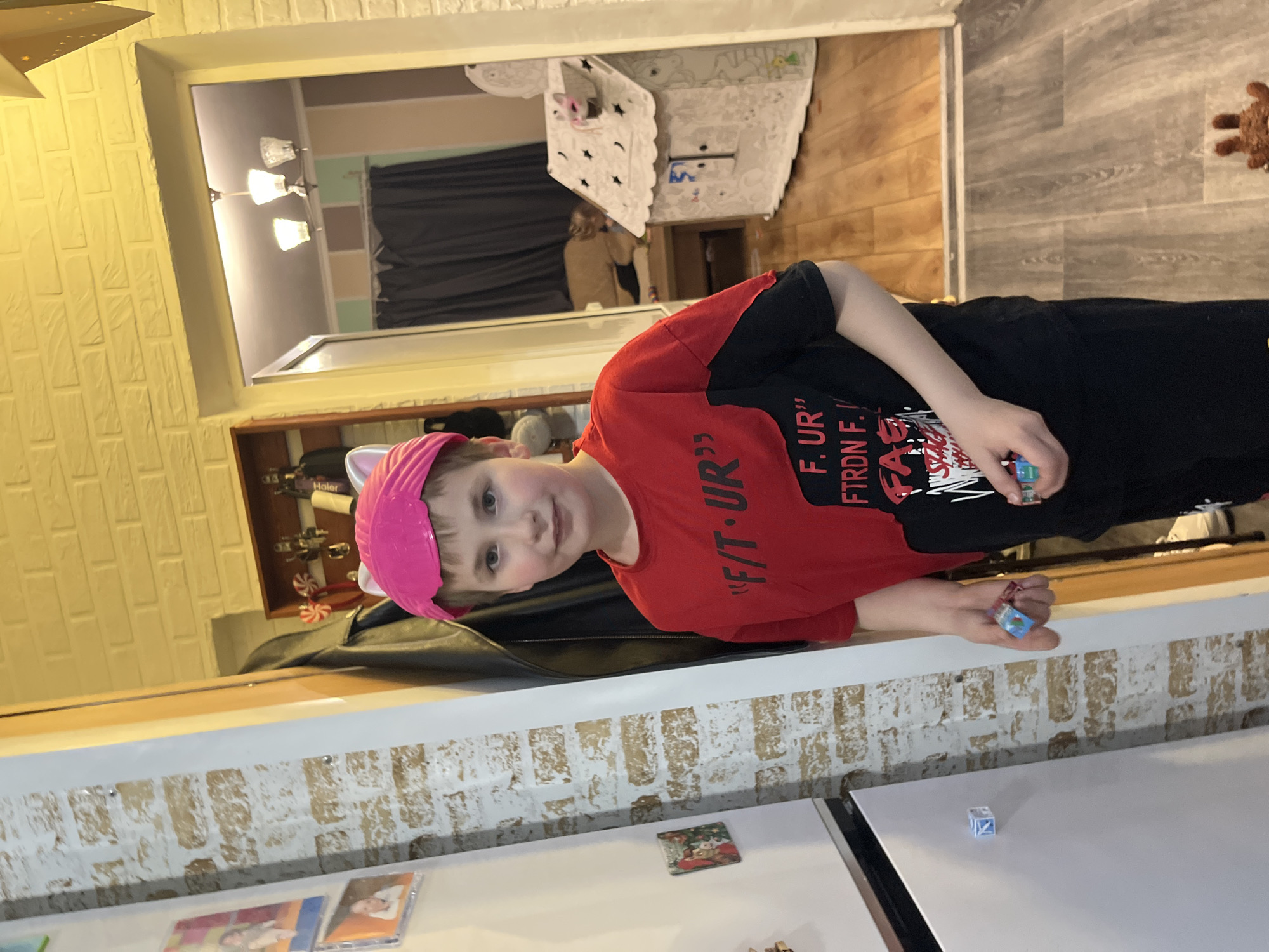 A young boy with fair skin and light-colored hair is wearing a pink headband with cat ears. He is also wearing a red t-shirt with black sleeves and black shorts. He is holding a pair of dice in his right hand. He is standing next to a white countertop with a brick-patterned wall behind it. There are magnets and photos on the refrigerator door. The countertop is cluttered. In the background, there is a glimpse of a room with a table covered with a patterned tablecloth and a person standing in the room. The location is not identifiable.