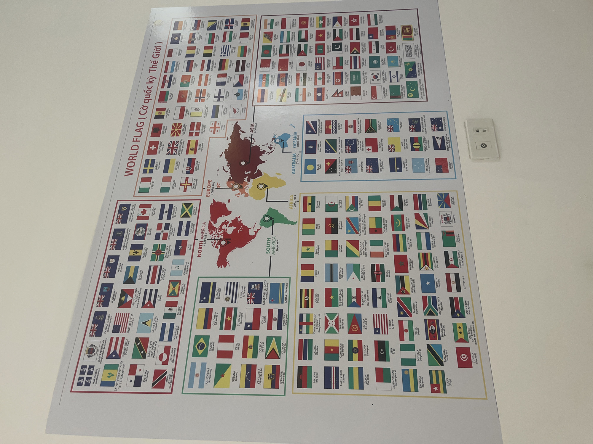 The image displays a large, colorful poster of the world map with flags of various countries categorized by continent. The poster is mounted on a white wall, with a light-colored electrical outlet visible on the lower right side of the wall. The poster is illuminated by overhead lighting. The text on the poster is in English and Vietnamese, indicating it is likely in a public space in Vietnam. The presence of the electrical outlet and the overall presentation suggest the poster is displayed indoors, possibly in an educational institution or a common area. Given the context of Đà Nẵng, Vietnam, this poster could be found in a school, a library, an airport, or a hotel lobby, serving as an educational or decorative piece. There are no people or specific actions depicted in the image.