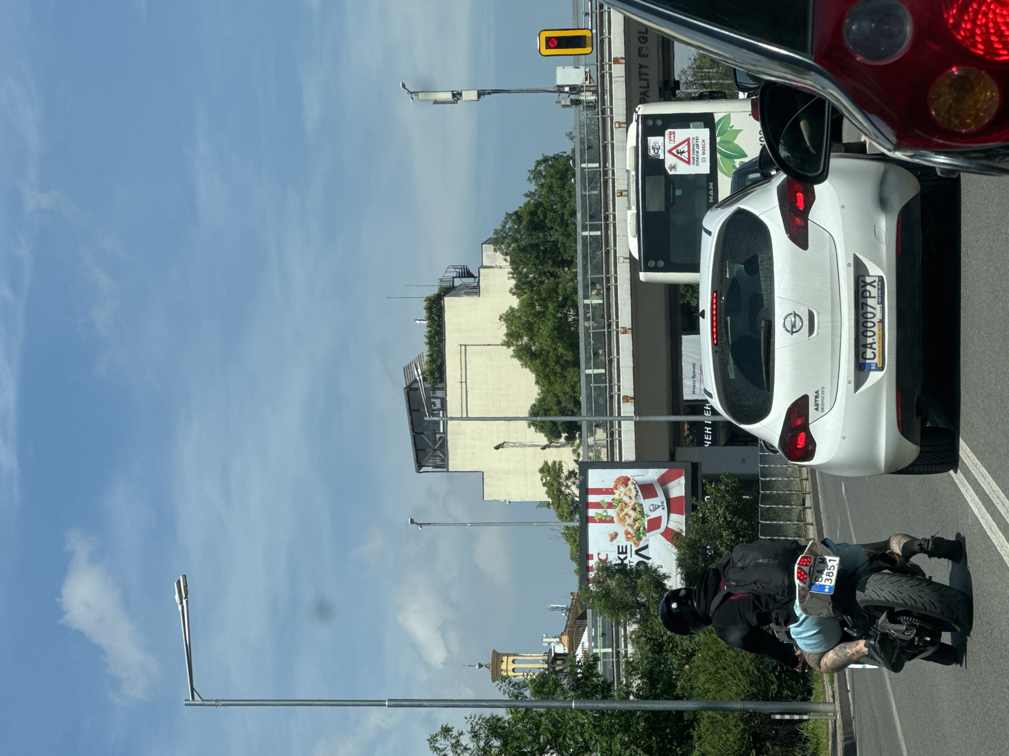 The image depicts a bright, sunny daytime street scene in Sofia, Bulgaria, taken from inside a vehicle, with part of its red taillight and side mirror visible in the upper right. The sky is clear blue with some wispy clouds.
In the immediate foreground, a white Opel Astra car (license plate CA.0007 PX) is partially visible. Behind it, a motorcyclist wearing a black helmet, black jacket, and light blue shorts is seated on a black motorcycle with license plate CA 3851 BM. Tattoos are visible on the rider's legs. Traffic appears to be stopped or moving slowly, possibly due to a red traffic light, which is visible overhead in the middle-right of the frame.
To the right of the white car, the front of a white MAN bus is seen. It displays a "WIFI" sticker, a green leaf logo, a Bosch logo, and a public safety message in Bulgarian: "УБИЙ СКОРОСТТА СПАСИ ДЕТЕ!" (Kill the speed, save a child!). Above the bus, part of a sign reads "PALITY GL".
In the mid-ground, a large billboard advertising KFC, featuring fried chicken, is prominent. Buildings and lush green trees line the street. Further in the background, a distinctive yellow church spire is visible amongst other buildings, adding a local architectural element. Streetlights and overhead infrastructure are also part of the urban landscape.
Other visible text includes "ASTRA ОЖЕВЕРЬС АУТО" (Ozhervels Auto) on the white car's license plate frame, and partially visible words on the KFC billboard like "KE", "ЧЕНІ ЕН", and "Имаш време!" (Do you have time!). The scene portrays a typical urban traffic environment.