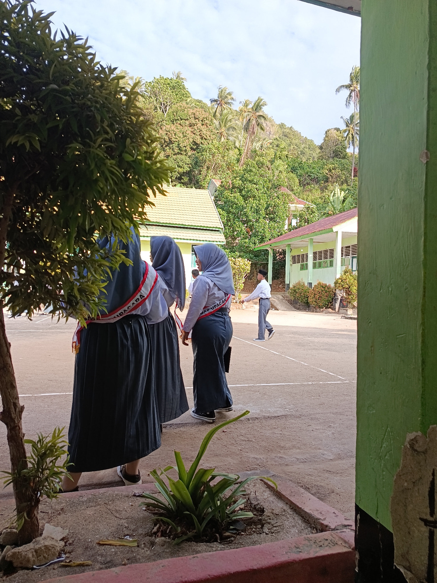 In the city of Mansalean, Indonesia, a sunny daytime scene unfolds in what appears to be a schoolyard. In the foreground, two students, identified as female by their attire, are seen from behind, wearing grey hijabs and long grey skirts. Over their white shirts, they wear red and white sashes, with the word "UPACARA" visible on one of them, suggesting a ceremonial occasion. To their right, another student, a male, dressed in a white shirt and grey pants, walks across the court. The background features a light green school building with a red-tiled roof, surrounded by lush green trees and palm trees, with a hilly landscape visible in the distance under a bright sky. The ground is paved with sand and a white line marks the boundary of the court. A large green pillar is prominently visible on the right side of the frame, partially obscuring the view of the school building.