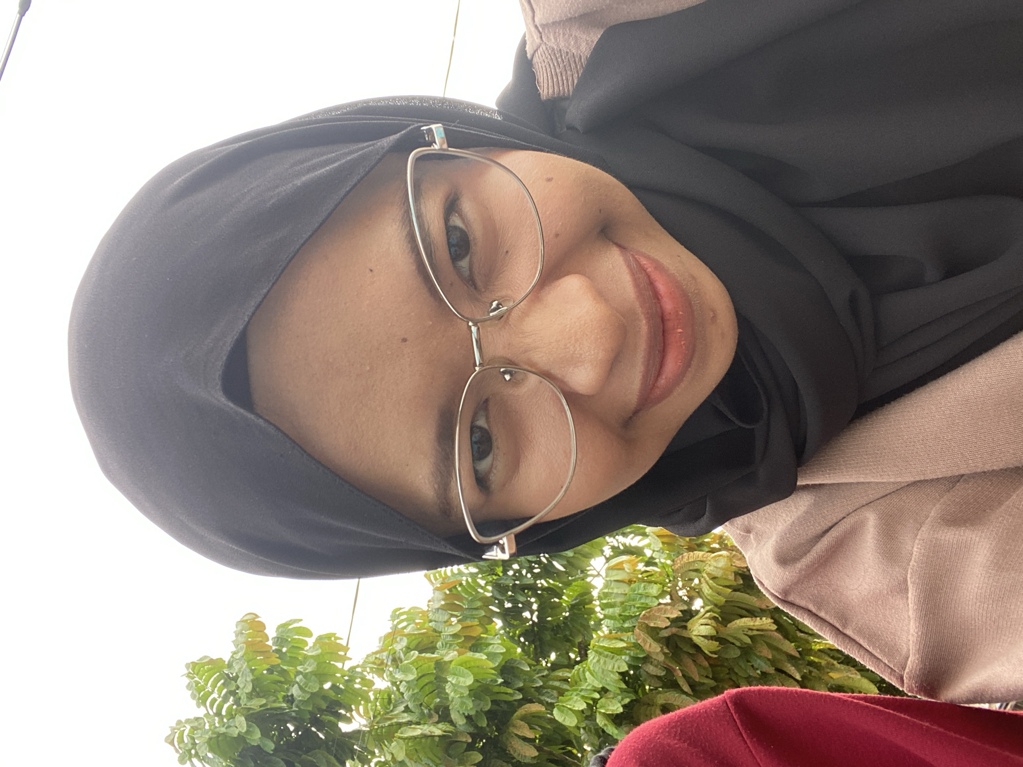 The media file is a selfie of a young woman wearing a black hijab and silver-rimmed glasses. She is smiling and looking directly at the camera. Her face is well-lit, suggesting it is daytime. She is wearing a light brown garment over a red garment, possibly a sweater or jacket. Lush green foliage is visible in the background, along with a bright, overcast sky. The image appears to have been taken outdoors, possibly in a garden or park. The overall mood is cheerful and relaxed. There is no visible text in the image. The location context is Air Molek, Indonesia.
