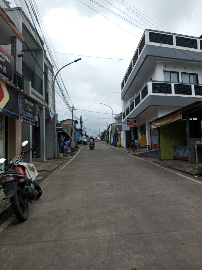 This image depicts a narrow concrete road stretching into the distance under an overcast sky, located in Melakudikadu, India.
On the immediate left, a black and orange motorcycle is parked, angled towards the road, partially on the sidewalk. Behind it, a multi-story building with a gray and white facade and dark balcony railings features a ground-level shop displaying a banner with "TARUM SUPER" text. Overhead, numerous black utility wires are densely strung between poles. Further down the left, additional buildings and a parked dark-colored car are visible.
On the immediate right, a three-story building, predominantly white with gray trim and dark, patterned balcony railings, has a ground-floor commercial space with a sign partially visible, displaying "AYAM KEPALA...". Adjacent to this building, a temporary structure with a bright yellow front and green side is visible, possibly a stall, near which two to three individuals are seated or standing.
In the middle of the concrete road, a single individual is operating a motorcycle, traveling away from the viewer. The road surface exhibits visible seams and minor irregularities. The sky is uniformly gray with no direct sunlight. No specific interactions between individuals are discernible.